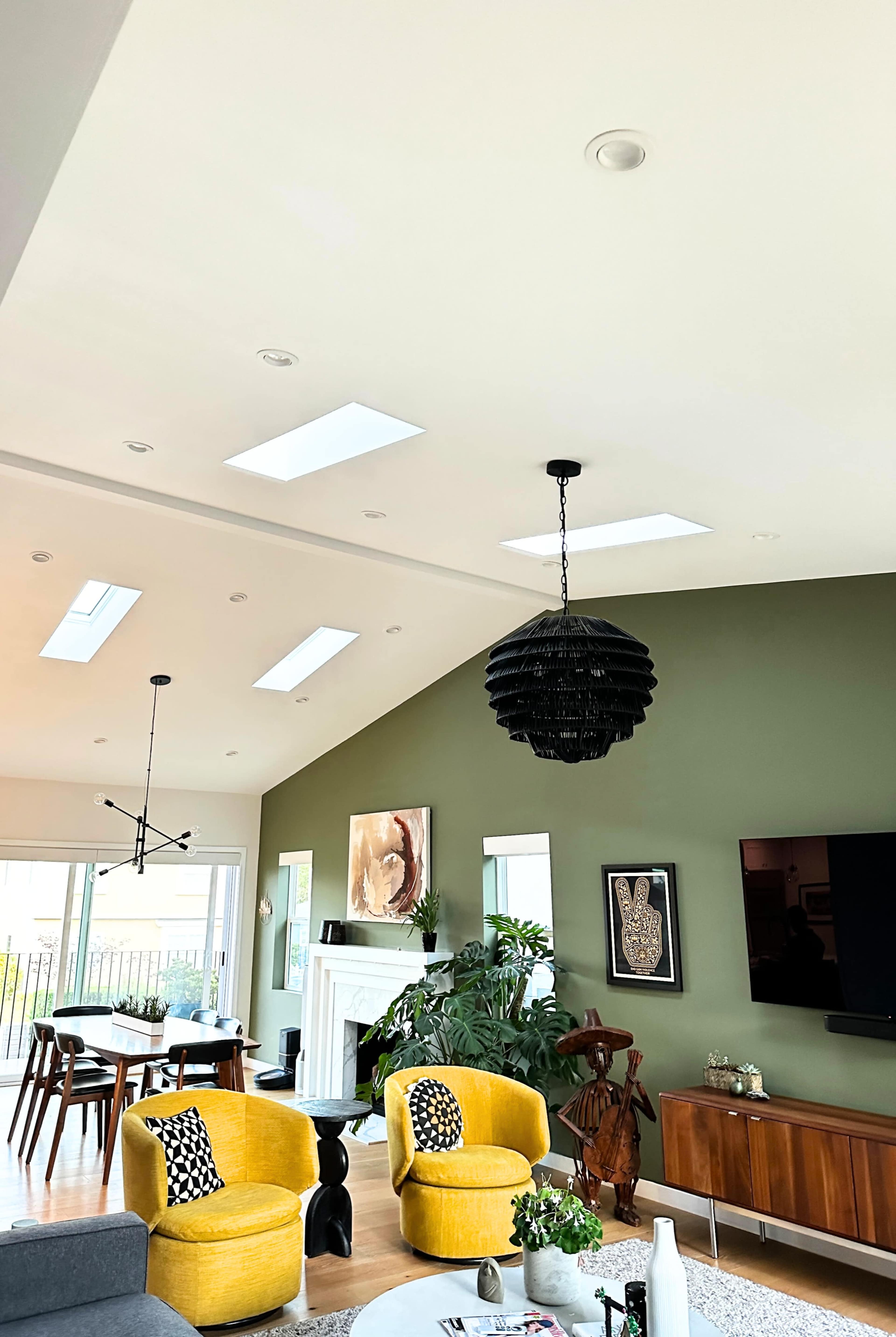 The image shows a modern living room with a green accent wall, two yellow chairs, a dark pendant light, and a mix of furniture including a dining table and a television cabinet.