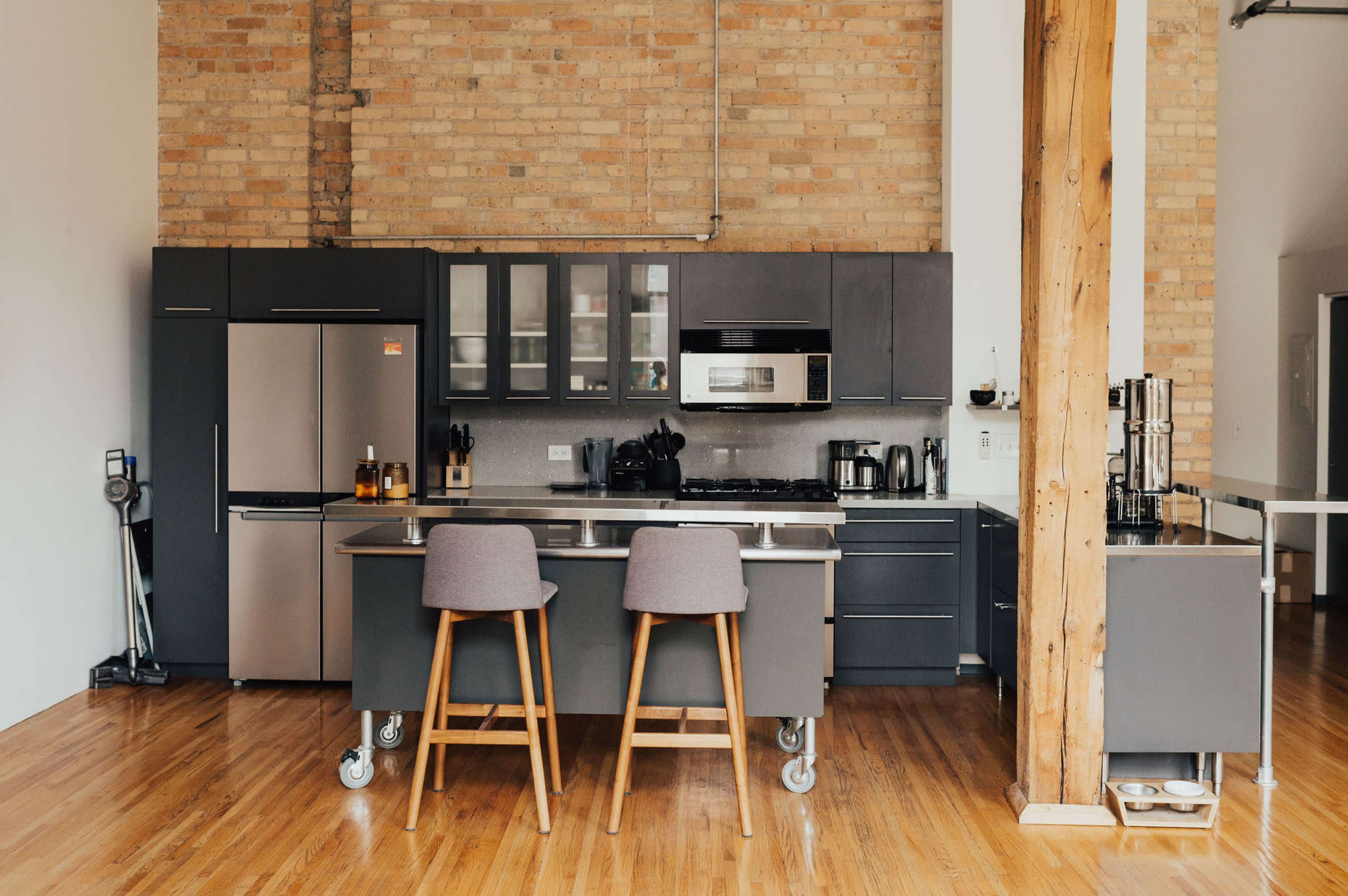 A modern kitchen with stainless steel appliances, dark cabinetry, and a central island with two bar stools, set against a backdrop of exposed brick walls.