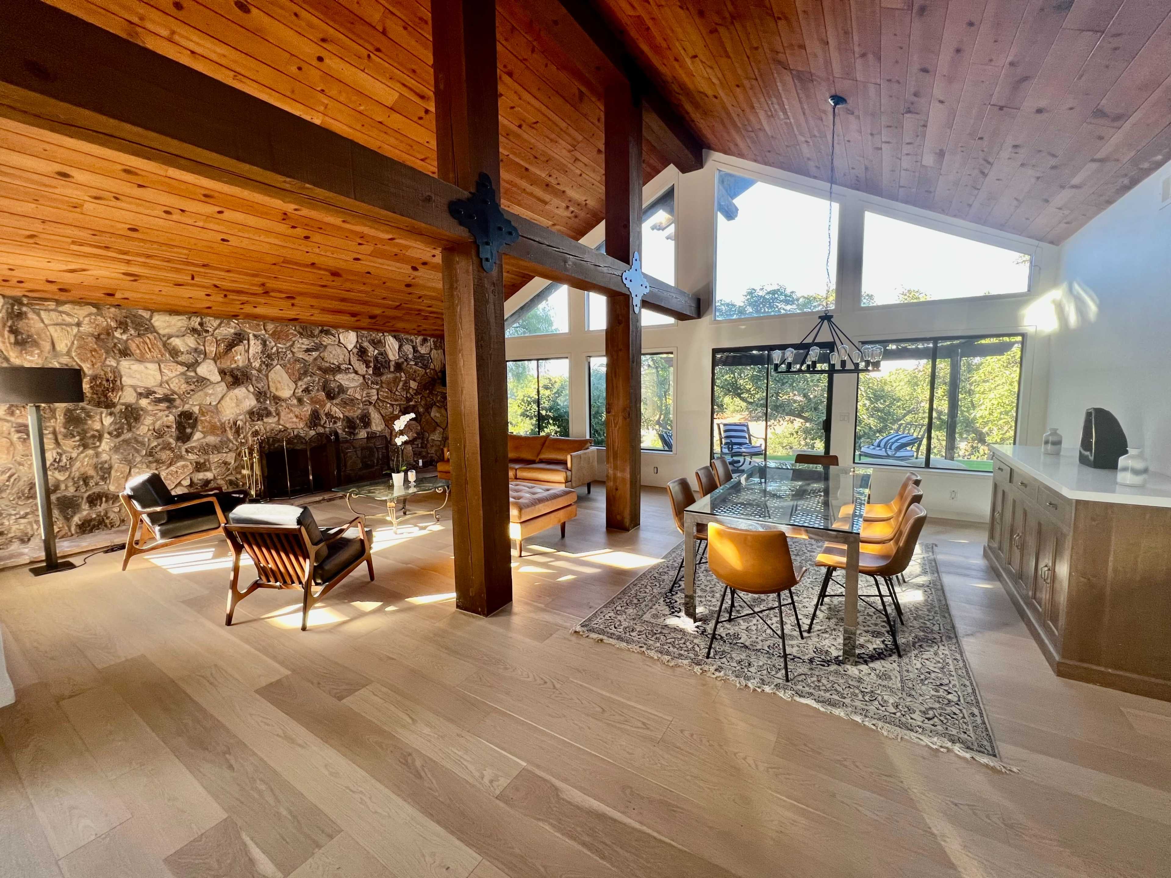 A modern living space with a wooden ceiling, stone wall, and a combination of seating areas and a dining table bathed in natural light from large windows.