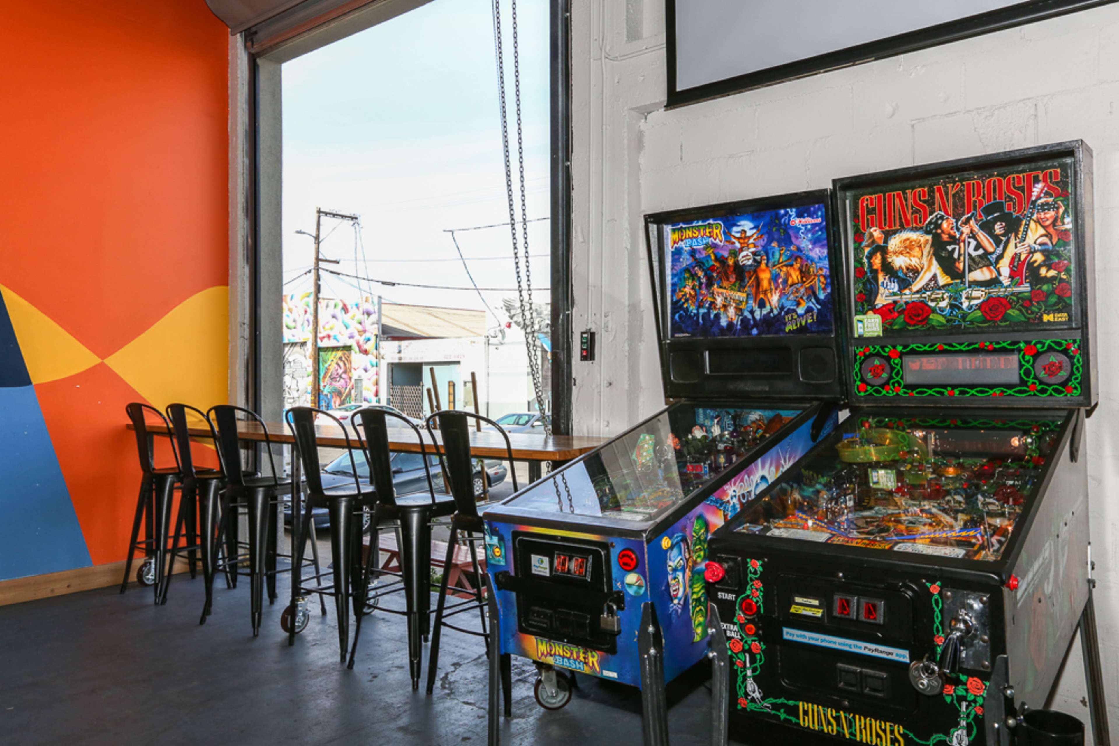 Two pinball machines beside a row of high-top bar stools in an industrial-style interior with large windows.