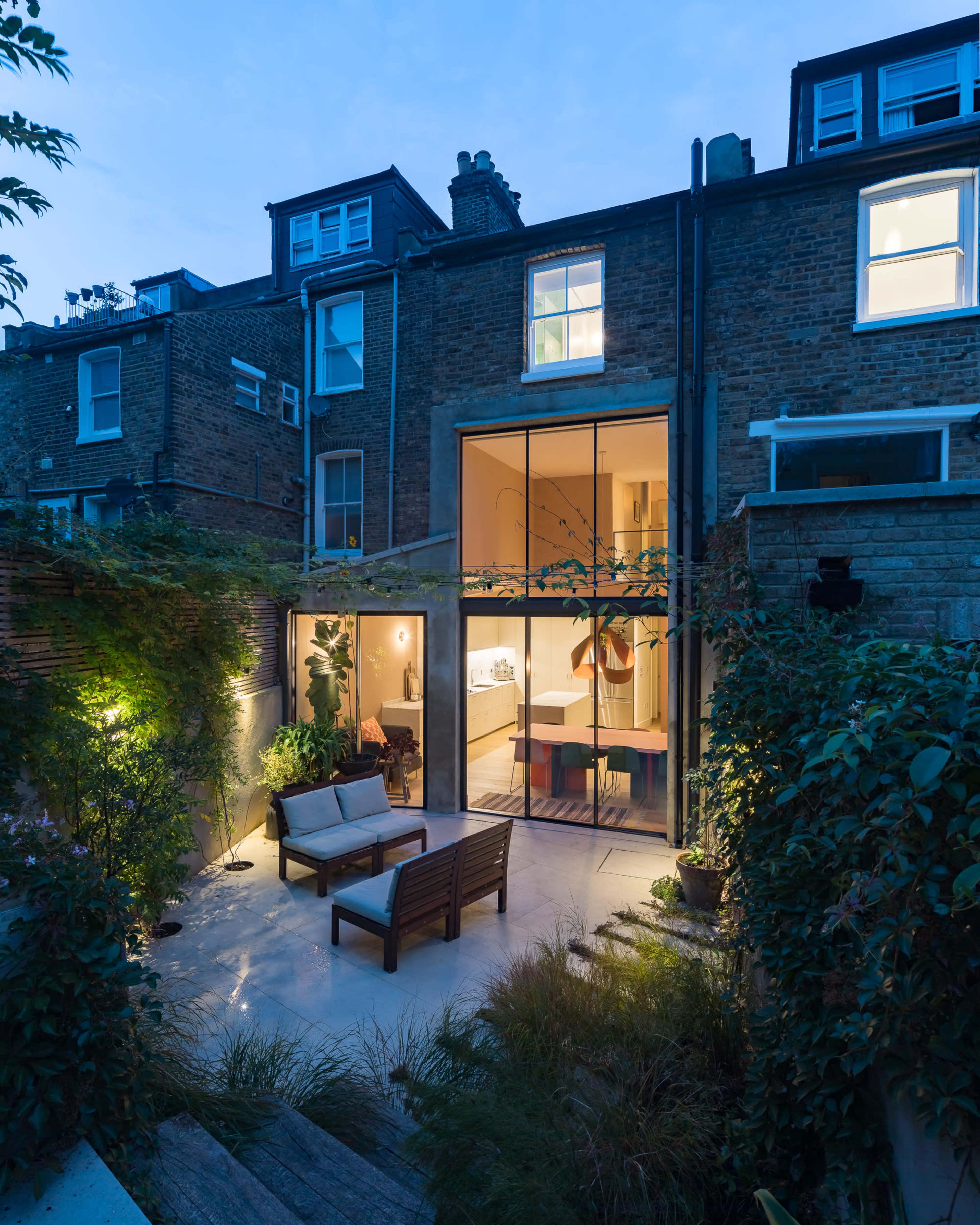 A modern backyard features a sleek, illuminated seating area surrounded by greenery, leading to a glass-walled kitchen and living space in an urban setting.
