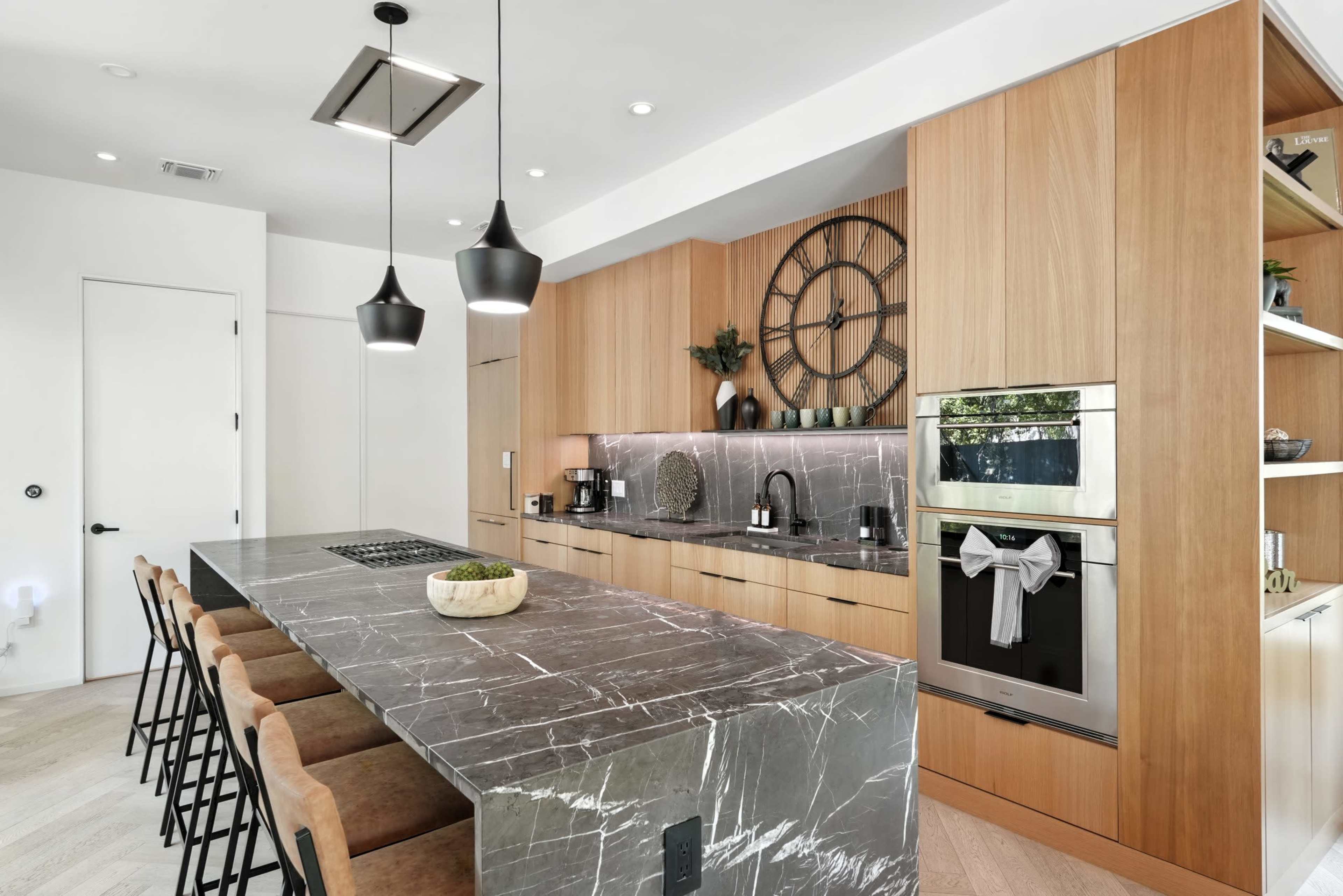A modern kitchen features a large marble island with bar stools, wooden cabinetry, and stainless steel appliances, complemented by stylish pendant lights.