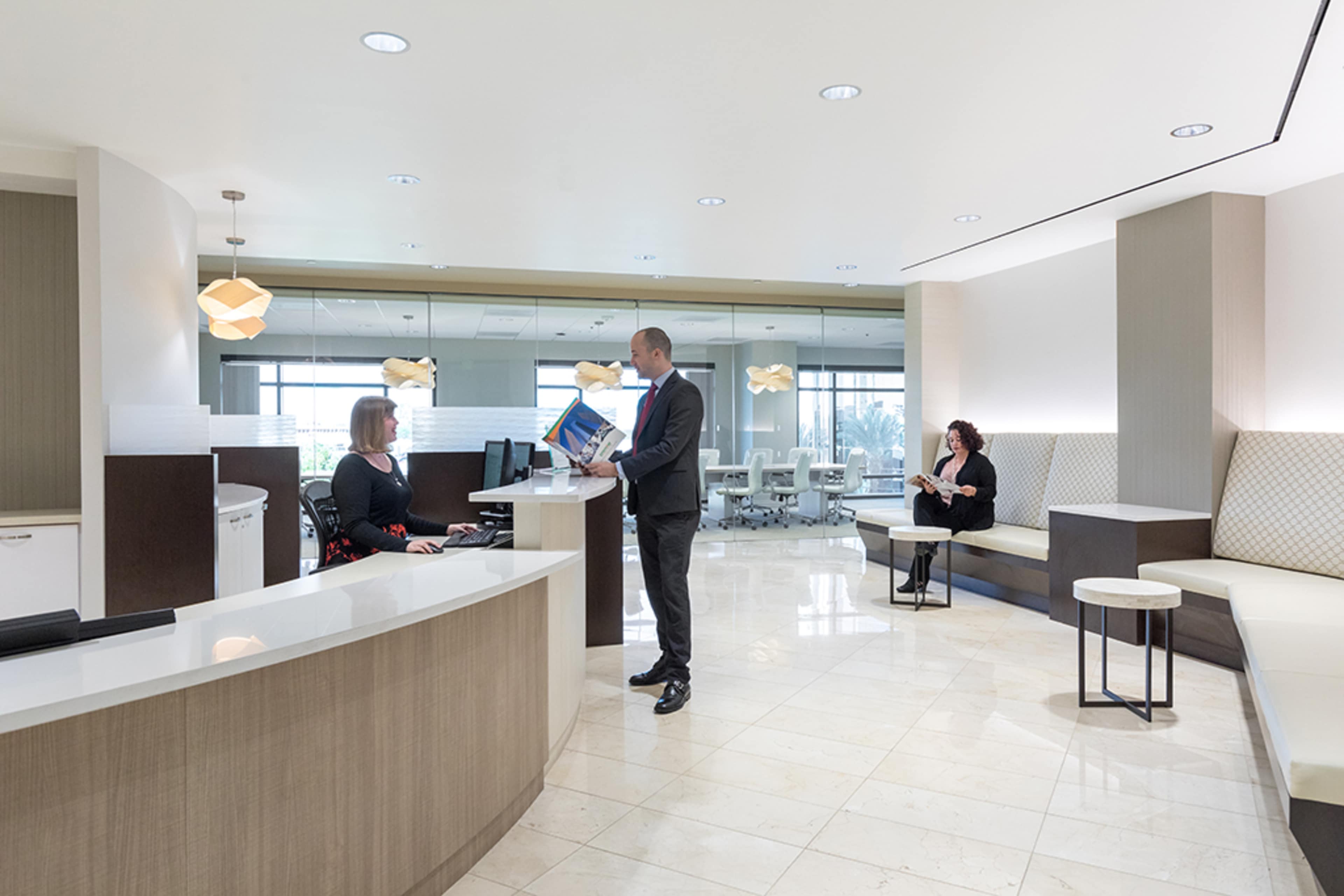 A man stands at a reception desk while a woman works on a computer, and another woman sits in a waiting area in a modern office lobby.