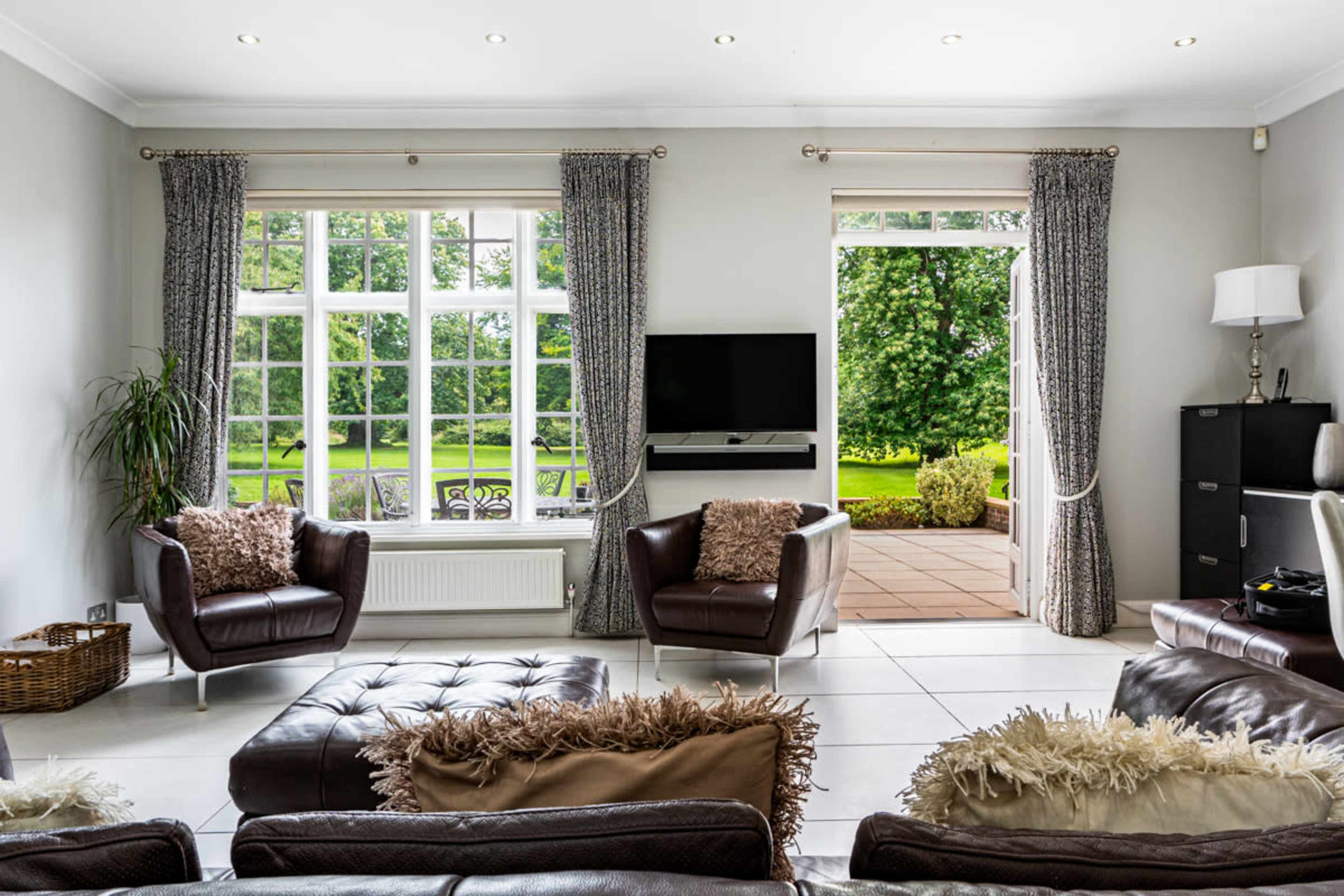 A modern living room with two leather armchairs, a television mounted on the wall, and a view of a patio and garden through large windows.