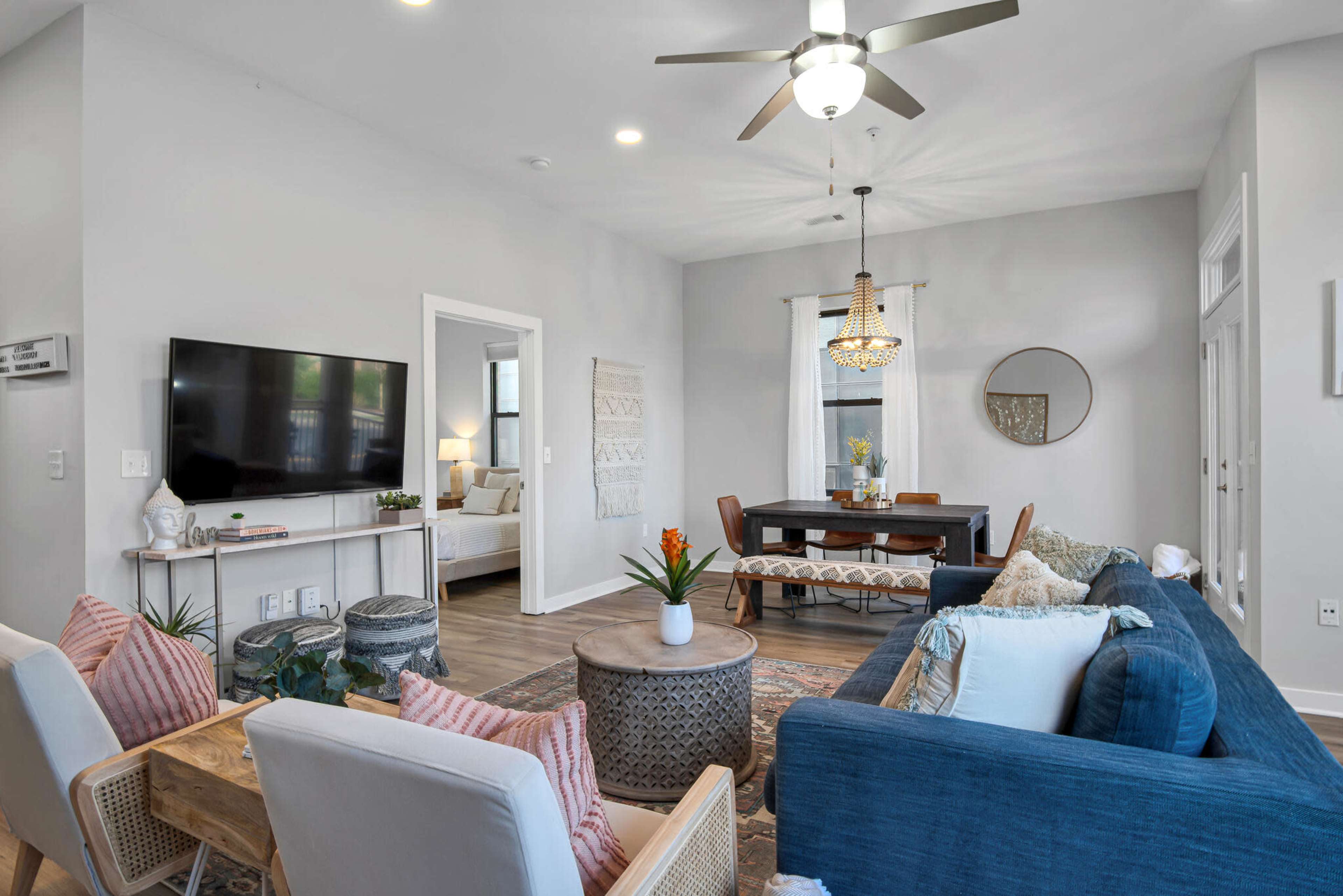 A modern living room features a blue couch, two armchairs, a coffee table, and a television, with a dining area visible through a doorway.