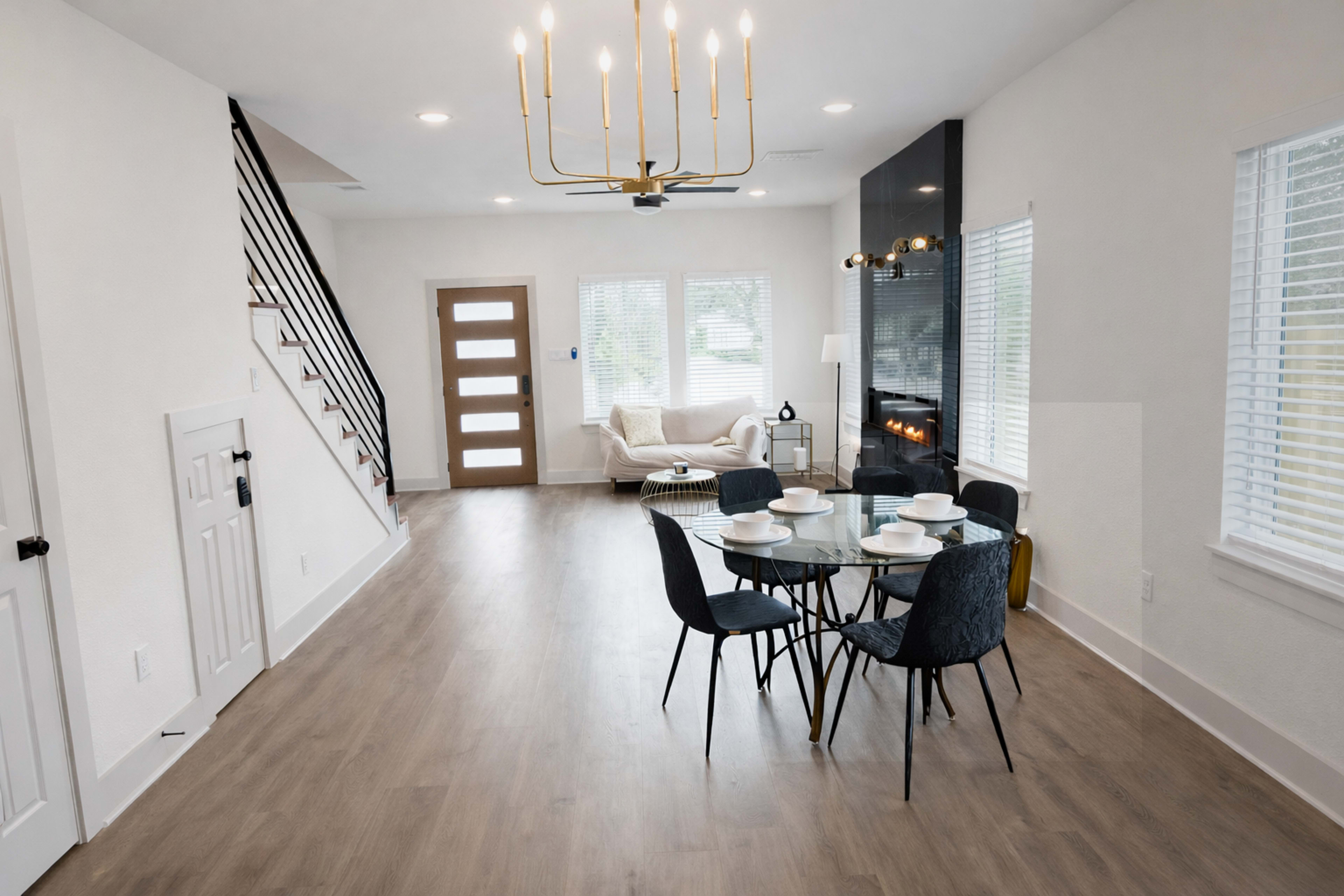 The image shows a modern, open-concept living space featuring a dining table set for four, a cozy seating area with a couch, and a staircase in the background.
