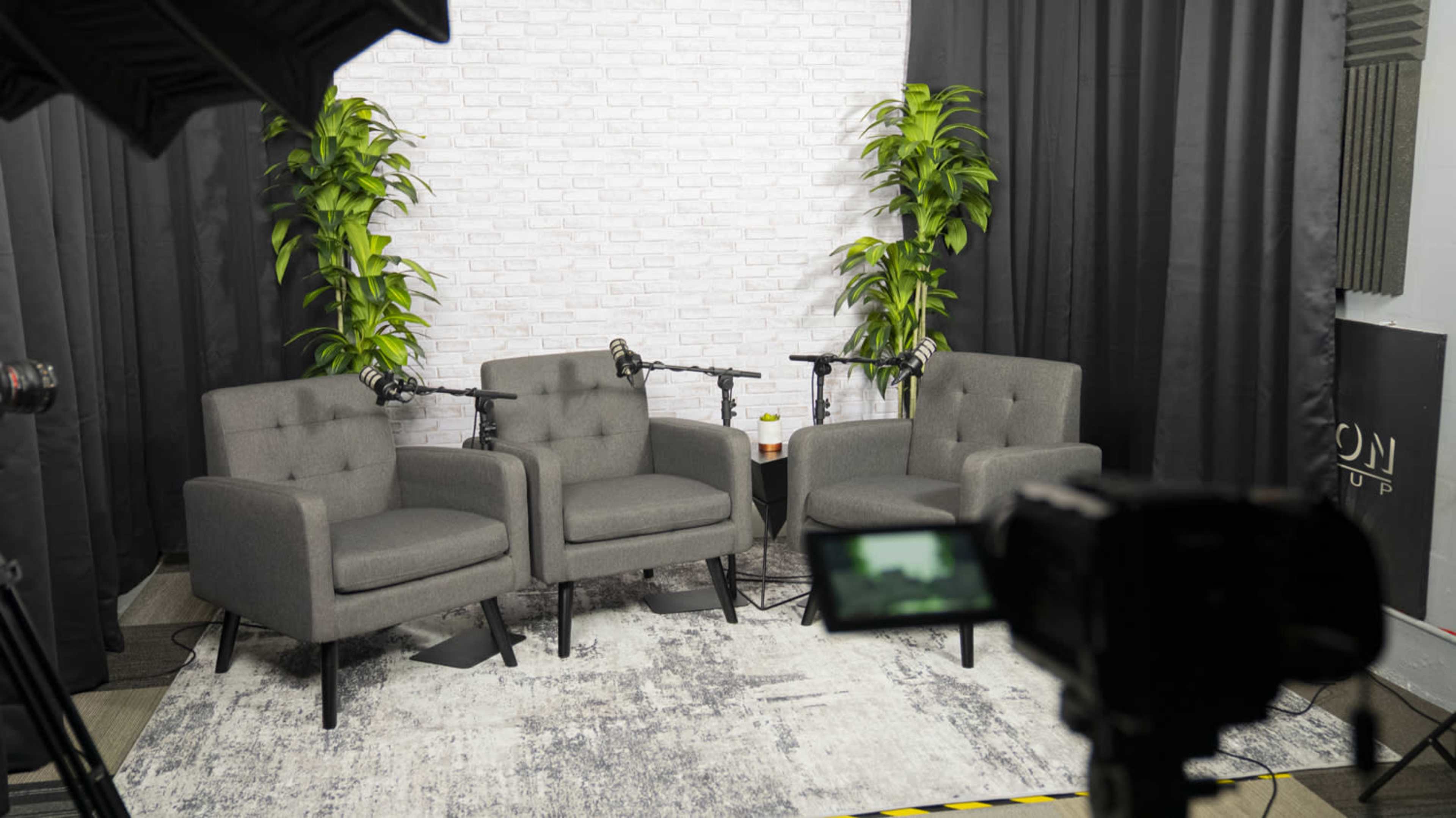 The image shows a set designed for a video recording, featuring three gray chairs with microphones on stands, potted plants, and a light-colored brick backdrop.
