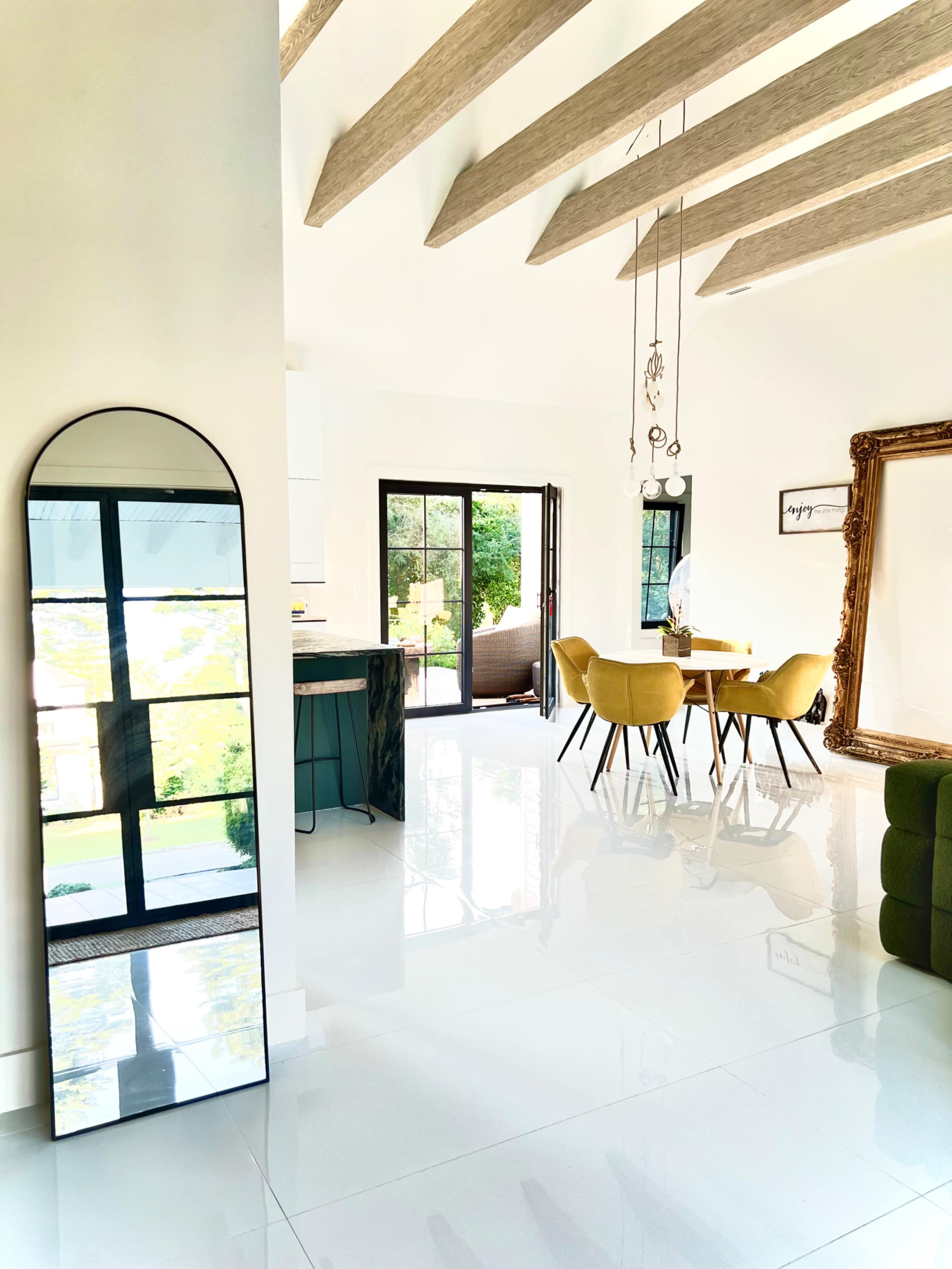 The image depicts a contemporary interior space featuring a large arched mirror, a dining area with yellow chairs, and large glass doors leading to an outdoor view.