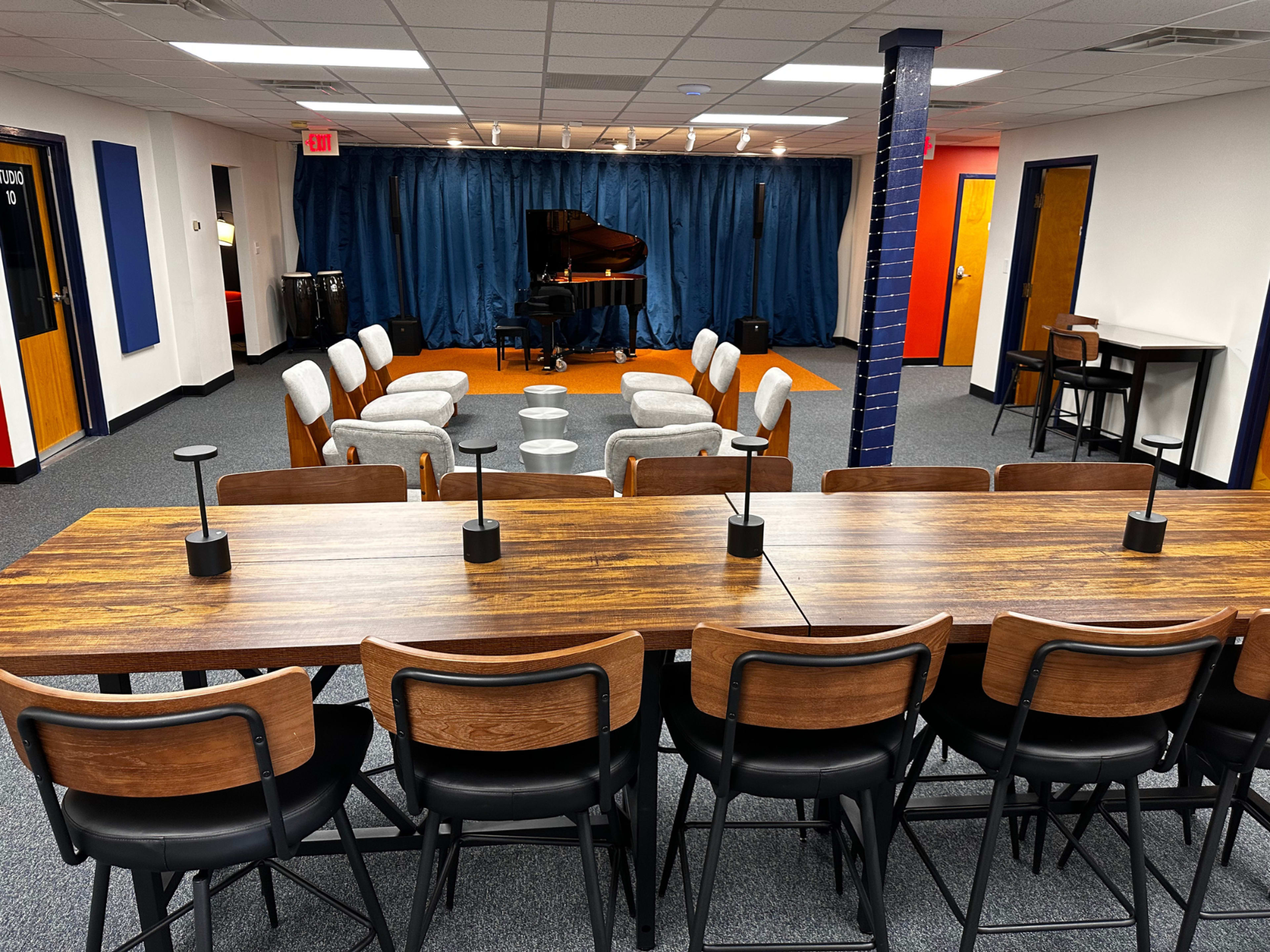 The image shows a room with a long wooden table and chairs, surrounded by a circular arrangement of light gray chairs, with a grand piano placed in the background against a blue curtain.