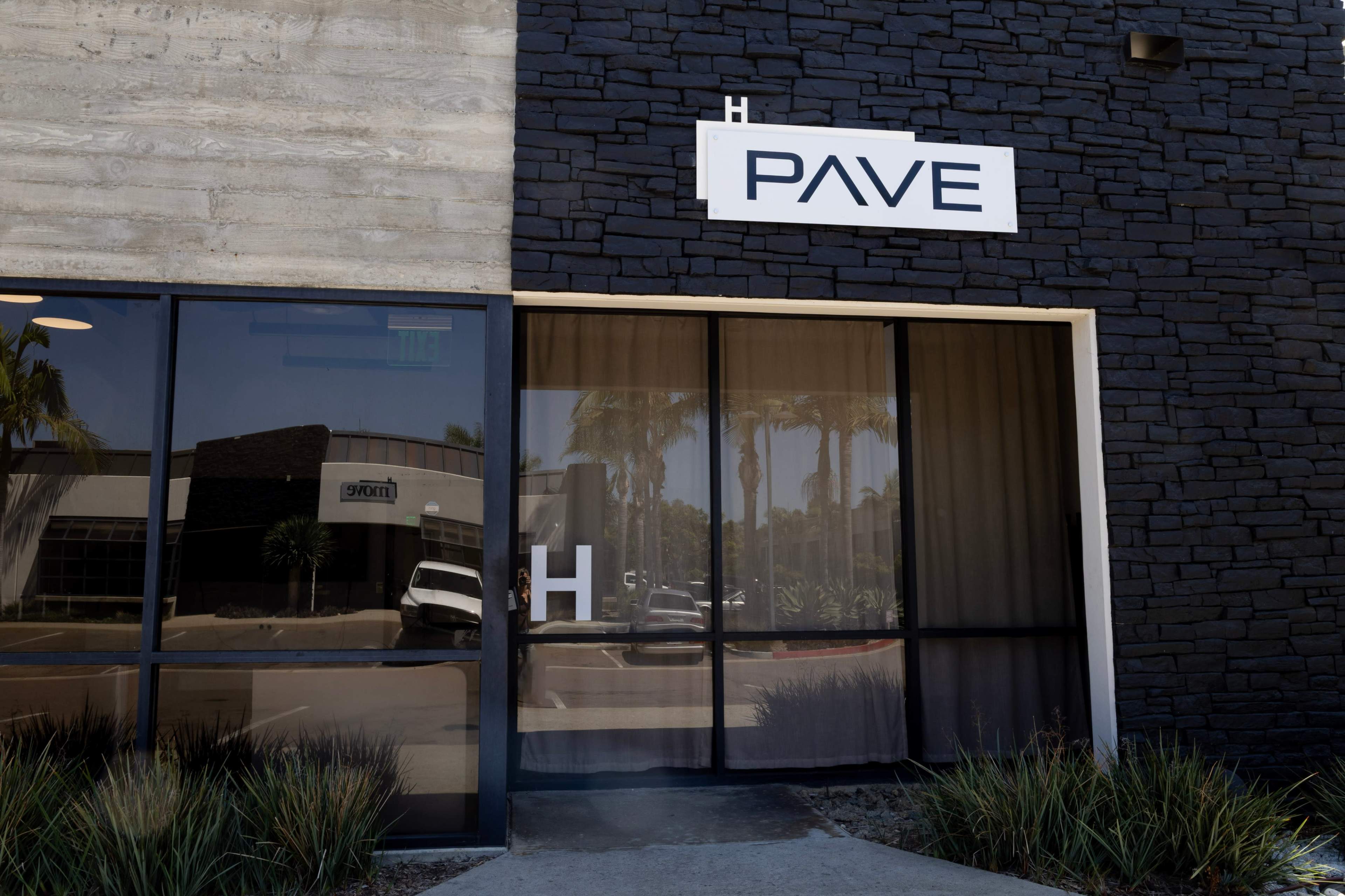 The image shows the exterior of a building with a modern design, featuring a large glass window displaying the name "PAVE" above an entrance marked with an "H."