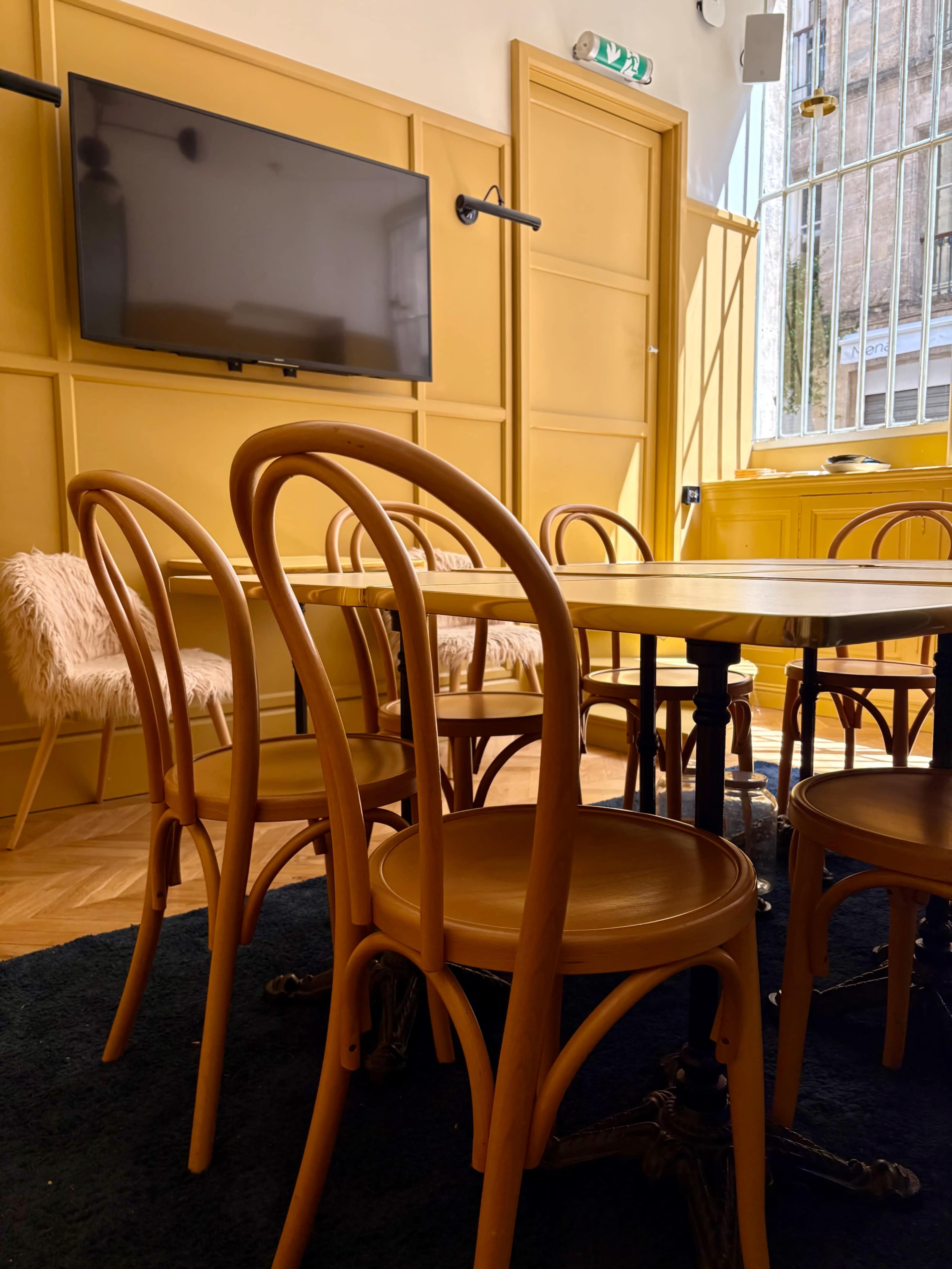 A cozy meeting room features a round wooden table surrounded by bentwood chairs, with a television mounted on the yellow walls.