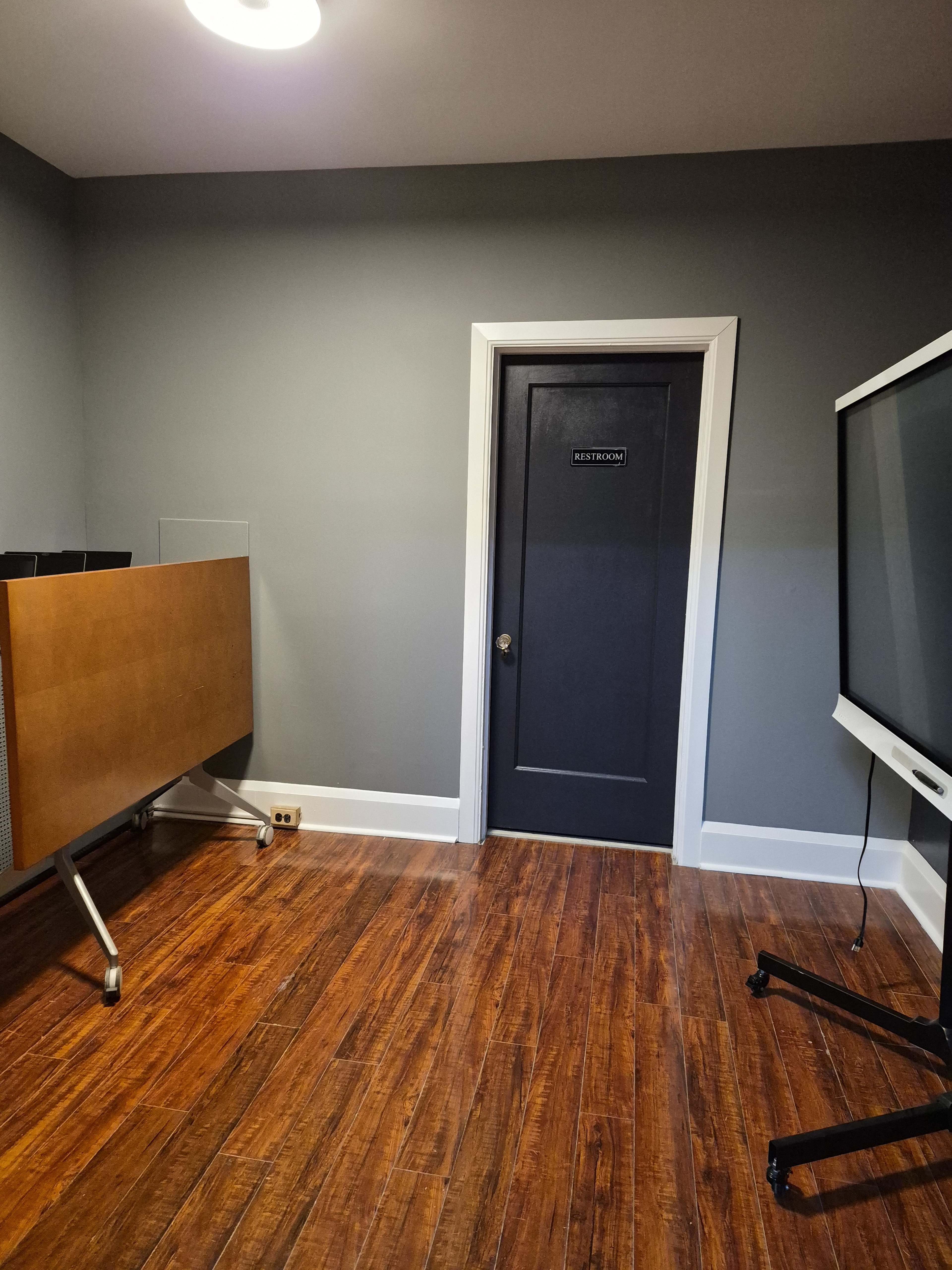 The image shows a room with dark gray walls, hardwood flooring, a wooden cabinet on wheels, a television on a stand, and a closed door labeled "RESTROOM."