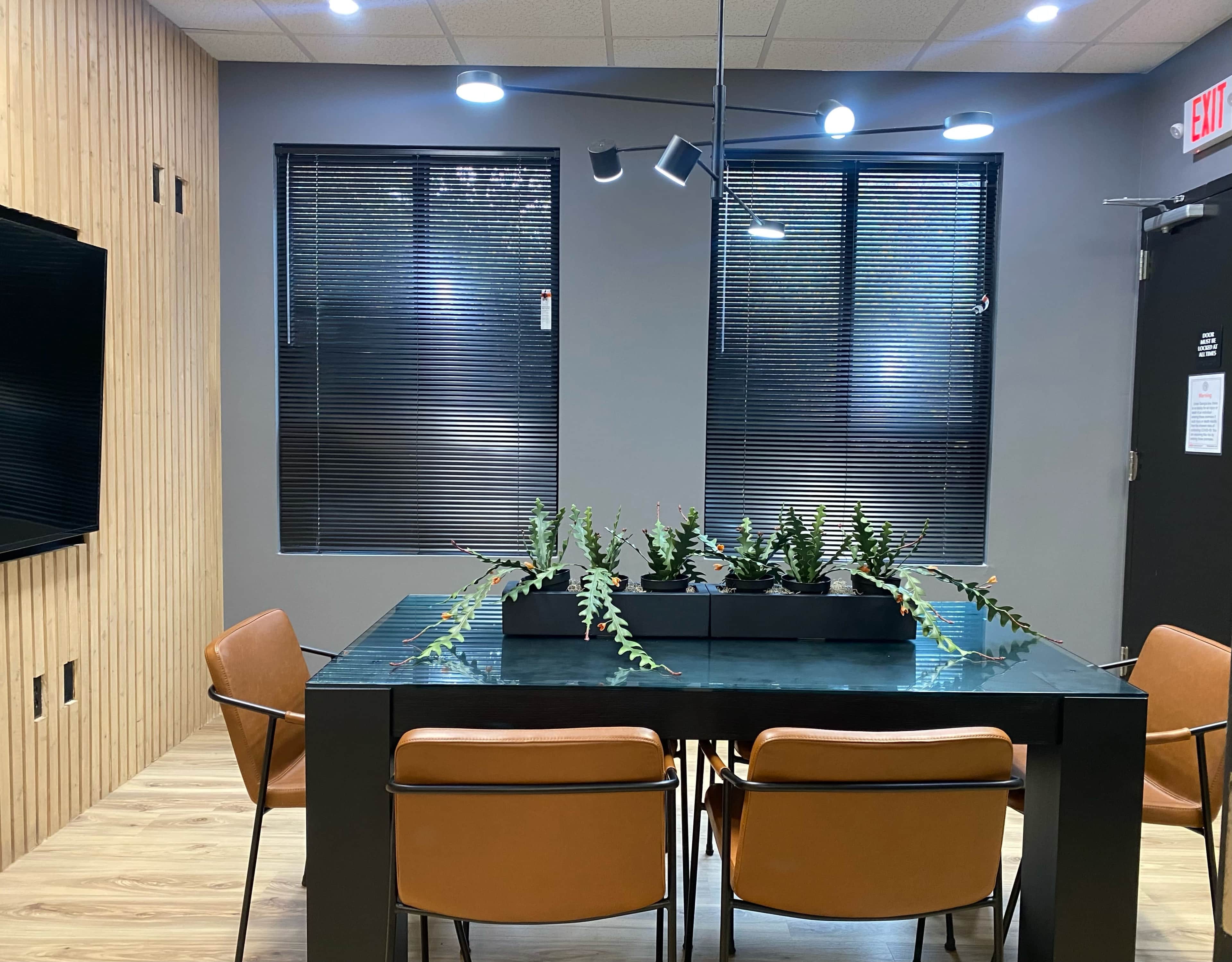 A modern conference room with a glass table surrounded by four brown chairs and a planter of green plants in the center, flanked by windows with blinds.