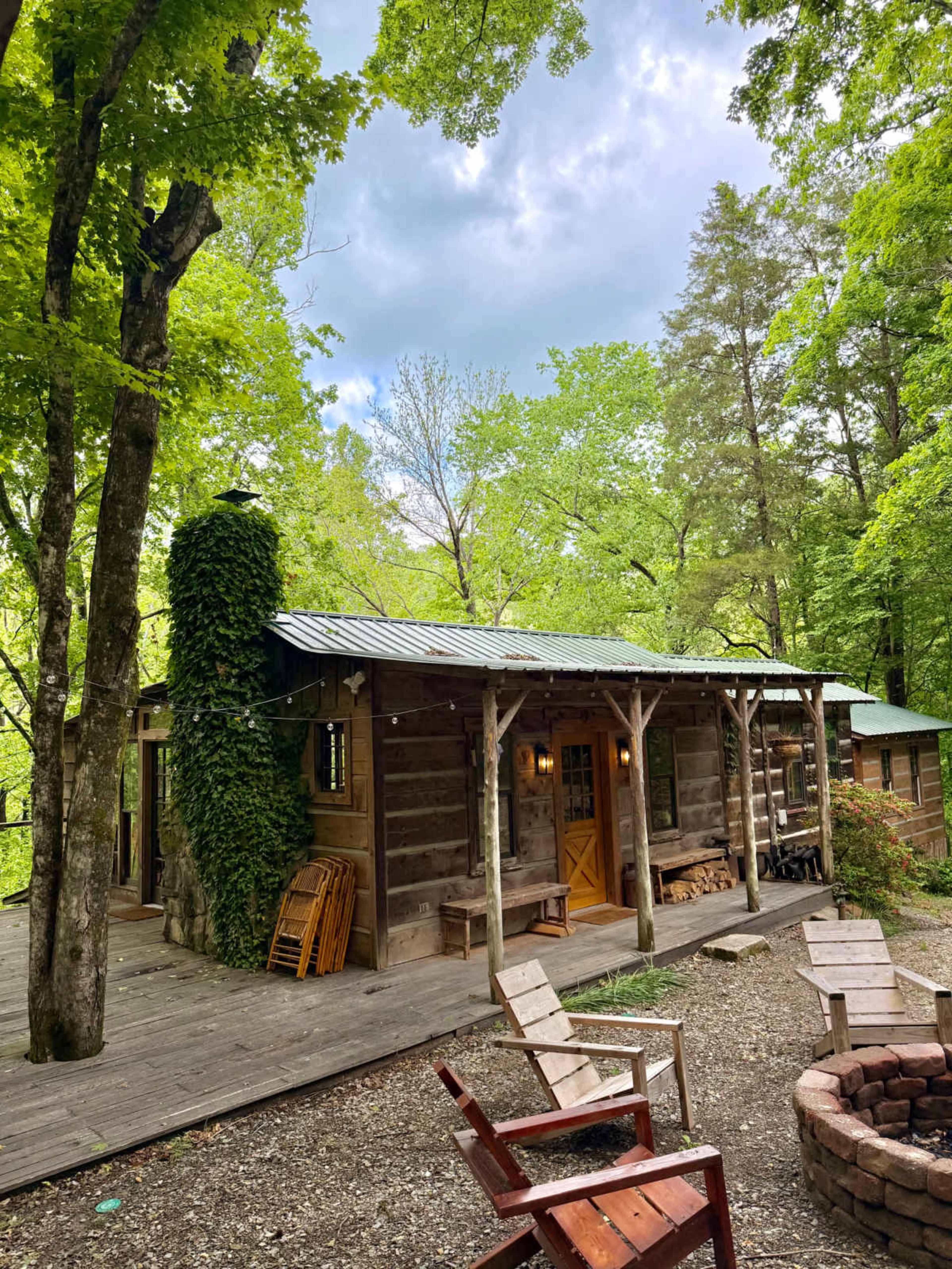 The image shows a rustic wooden cabin surrounded by lush green trees, with a wooden deck and seating area in the foreground.