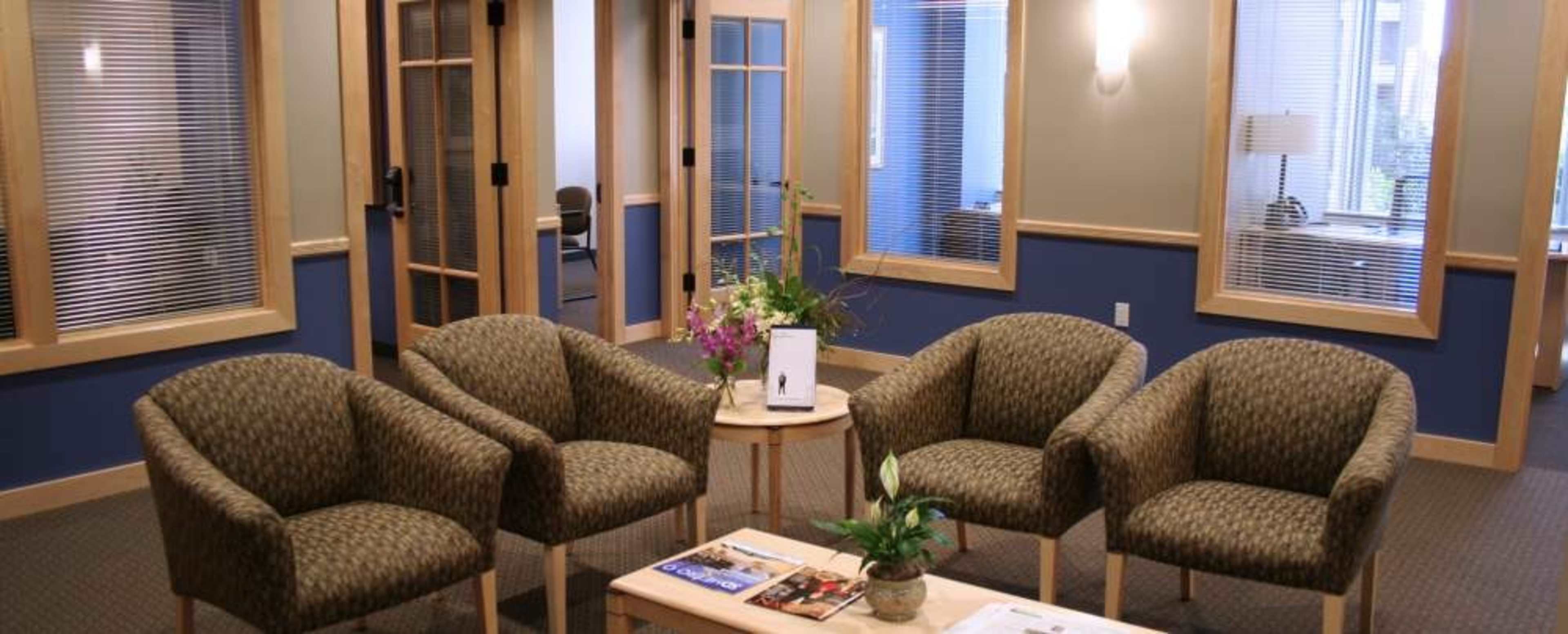 A waiting area with four patterned chairs arranged around a small wooden table, surrounded by glass-panel doors leading to adjacent rooms.