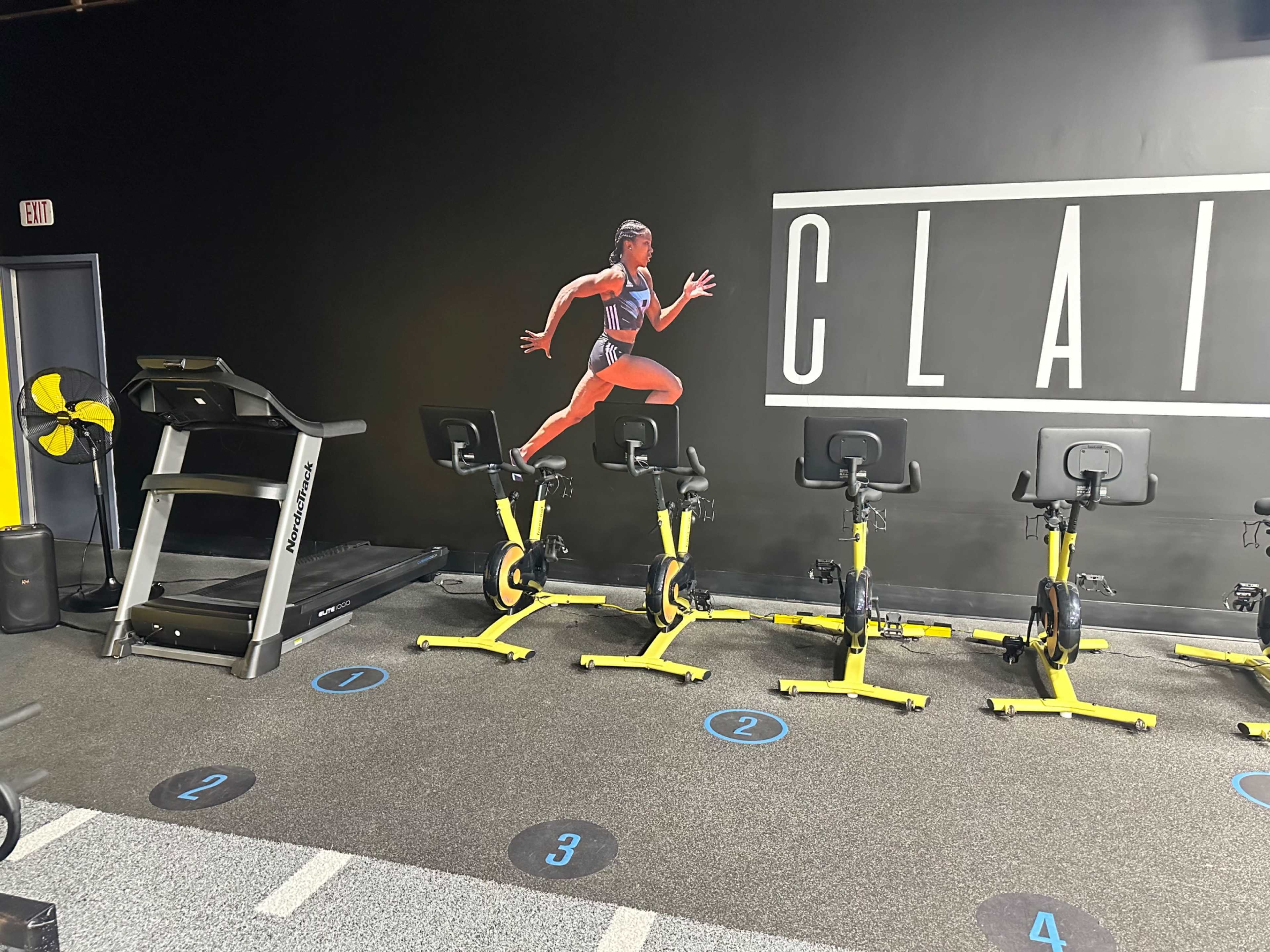 A gym area featuring a treadmill and several stationary bikes arranged on a dark wall with a large poster of a runner.