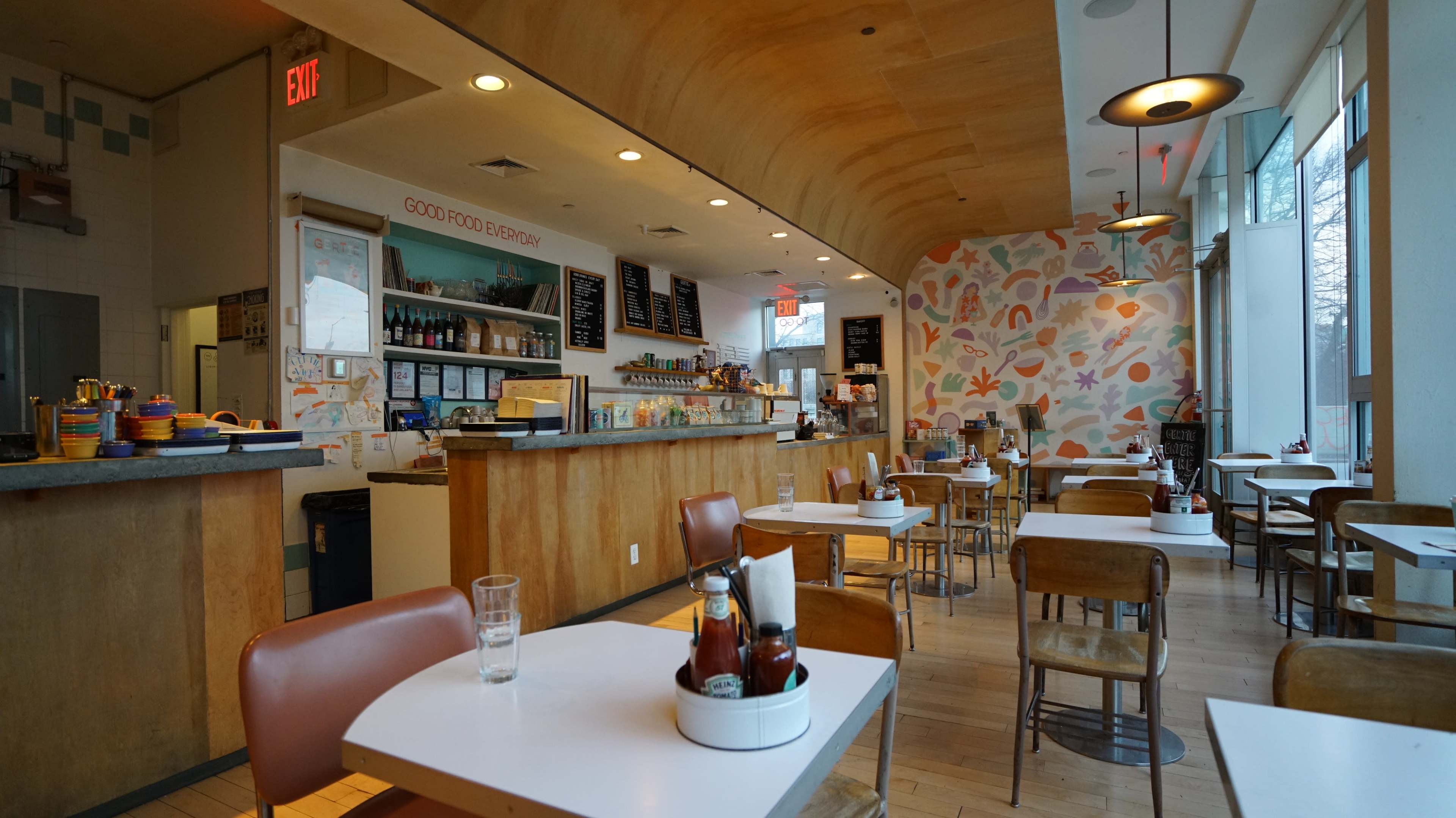 The image shows a brightly lit café interior with tables and chairs arranged around a service counter decorated with colorful artwork on the walls.