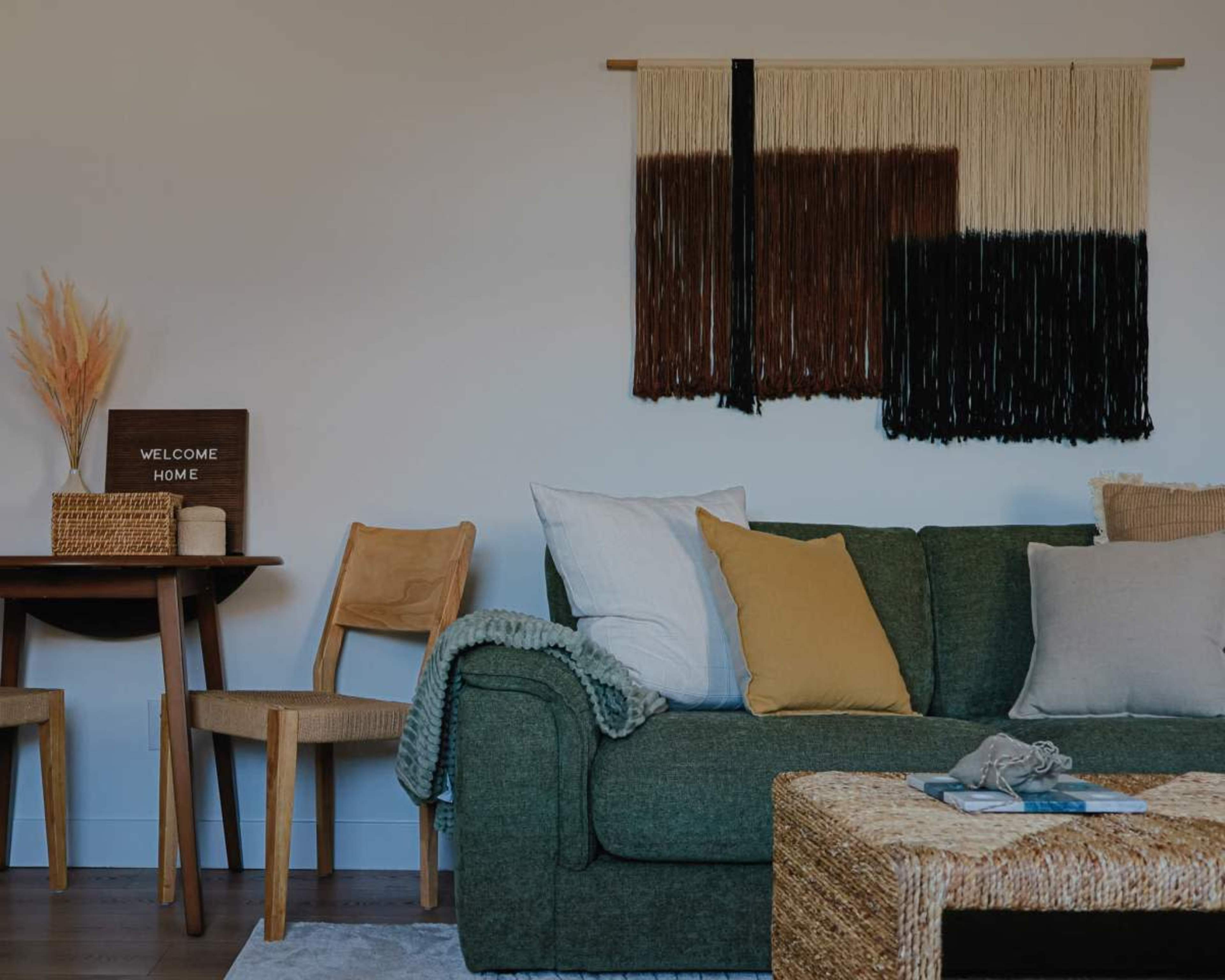A green couch with various pillows sits in a room next to a wooden table with a decorative welcome sign and a woven basket.