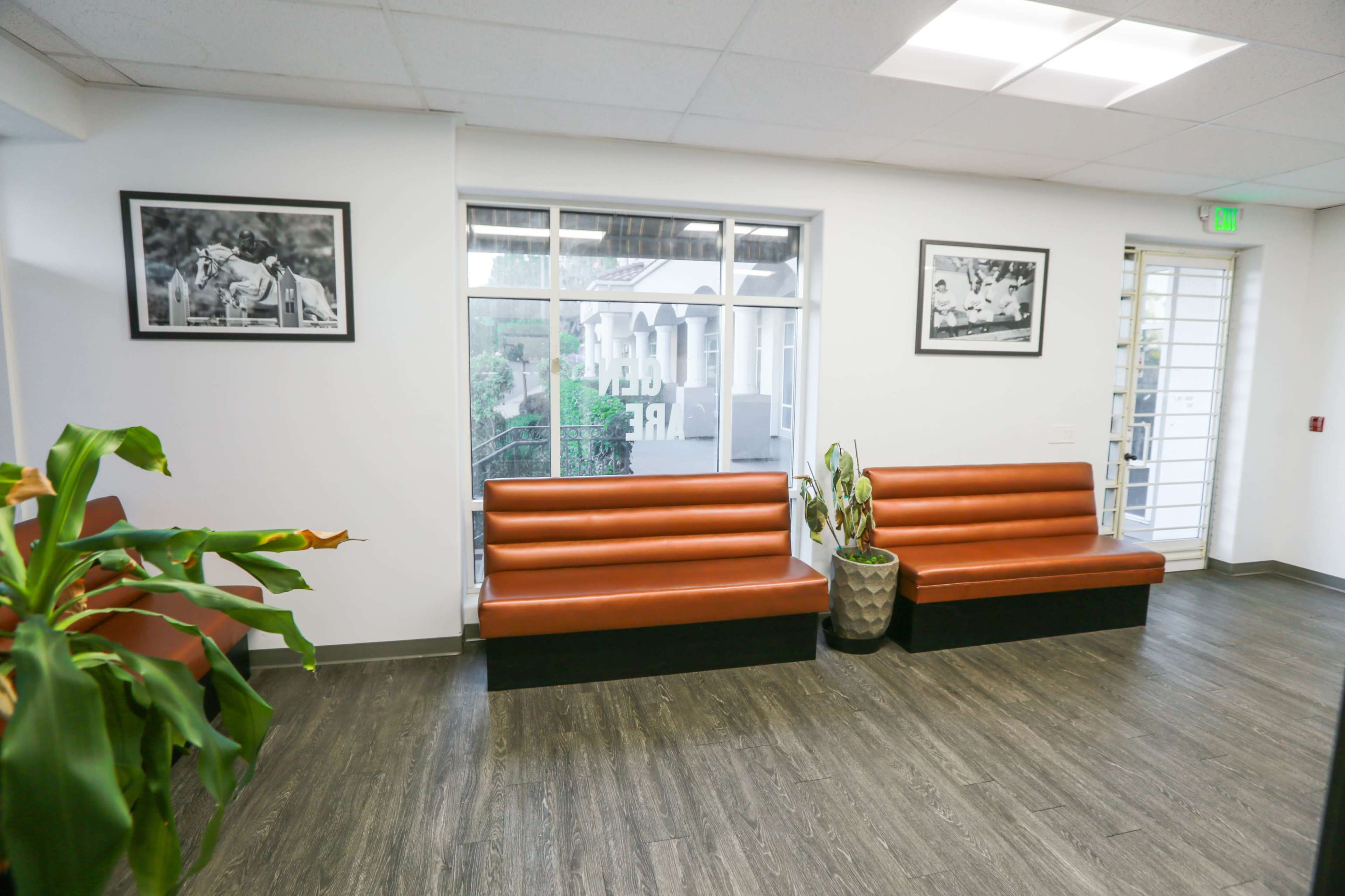 The image shows a modern waiting area with two brown benches, a large window, and potted plants.