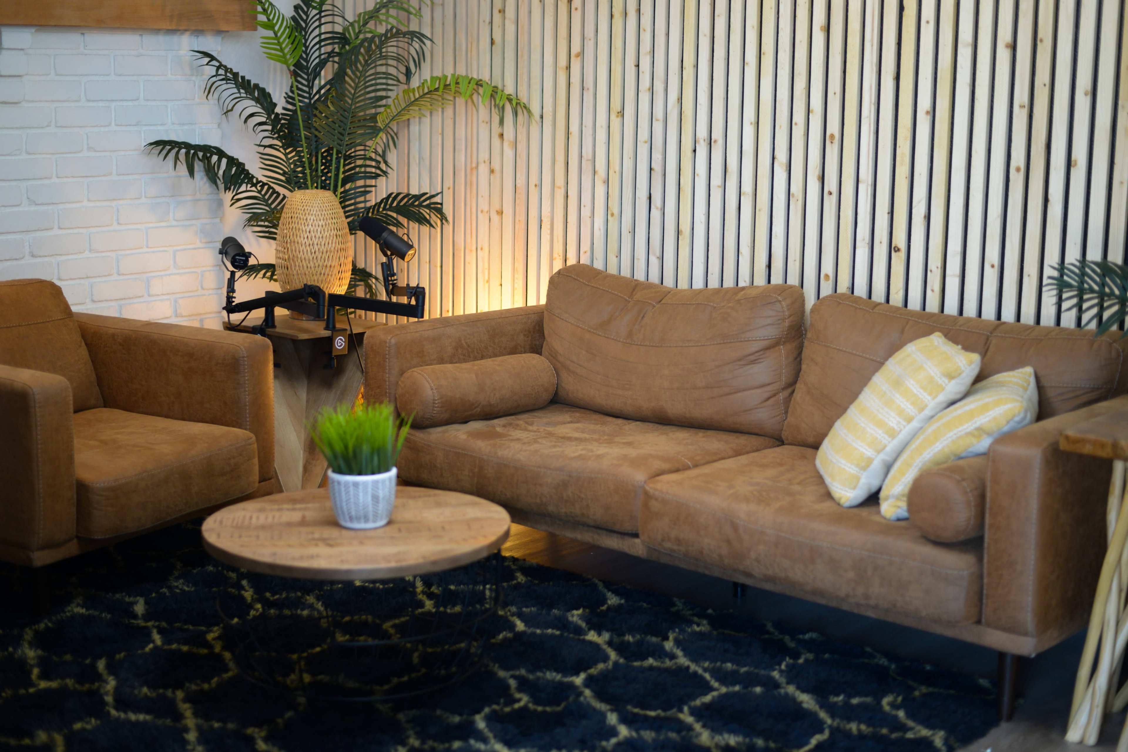 The scene features a stylish living room with a brown leather sofa, an armchair, a round wooden coffee table, and a potted plant against a wall of vertical wooden slats.