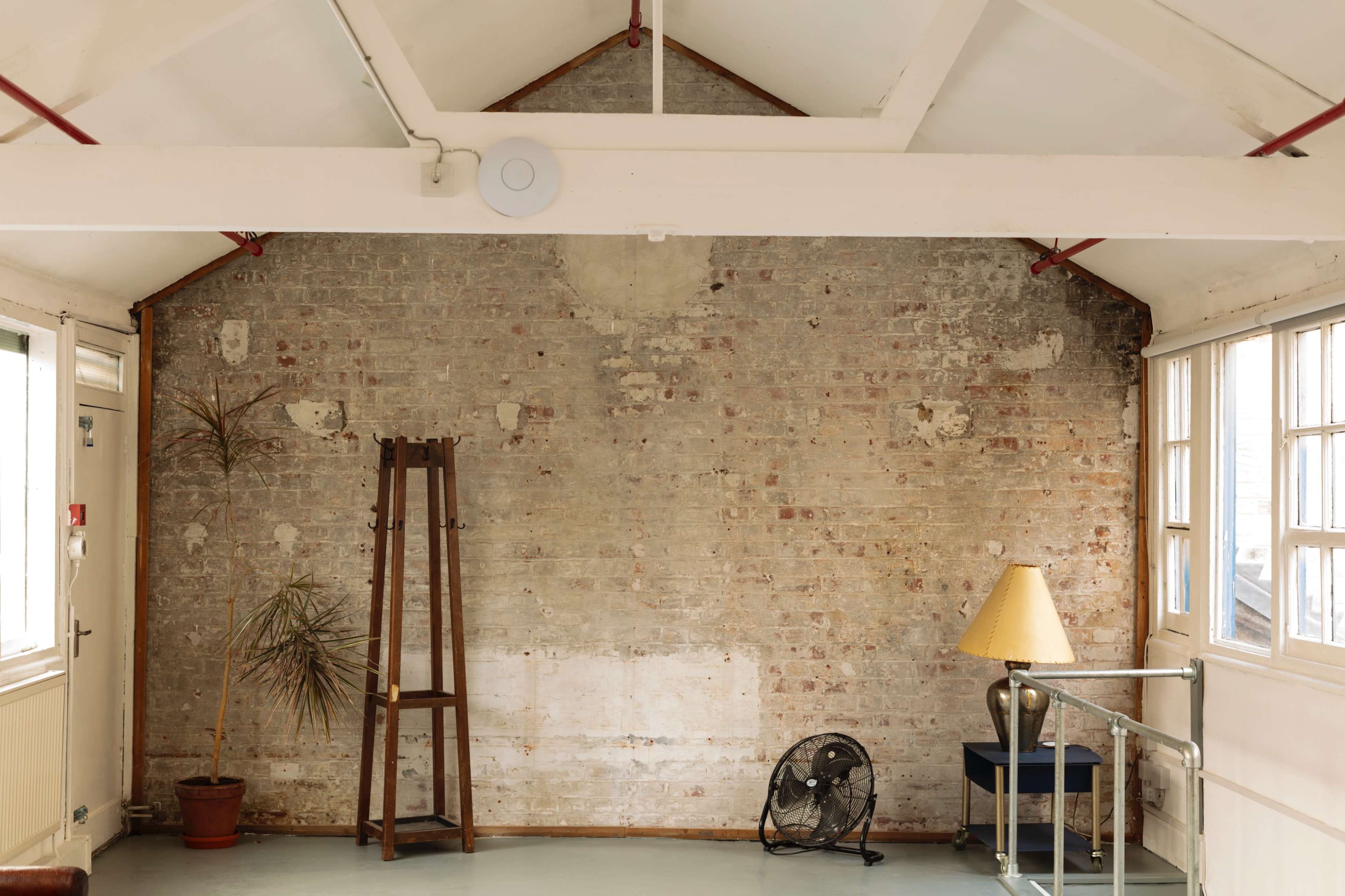 The image shows a room with a brick wall, a potted plant, a wooden ladder, a floor lamp, and a fan.