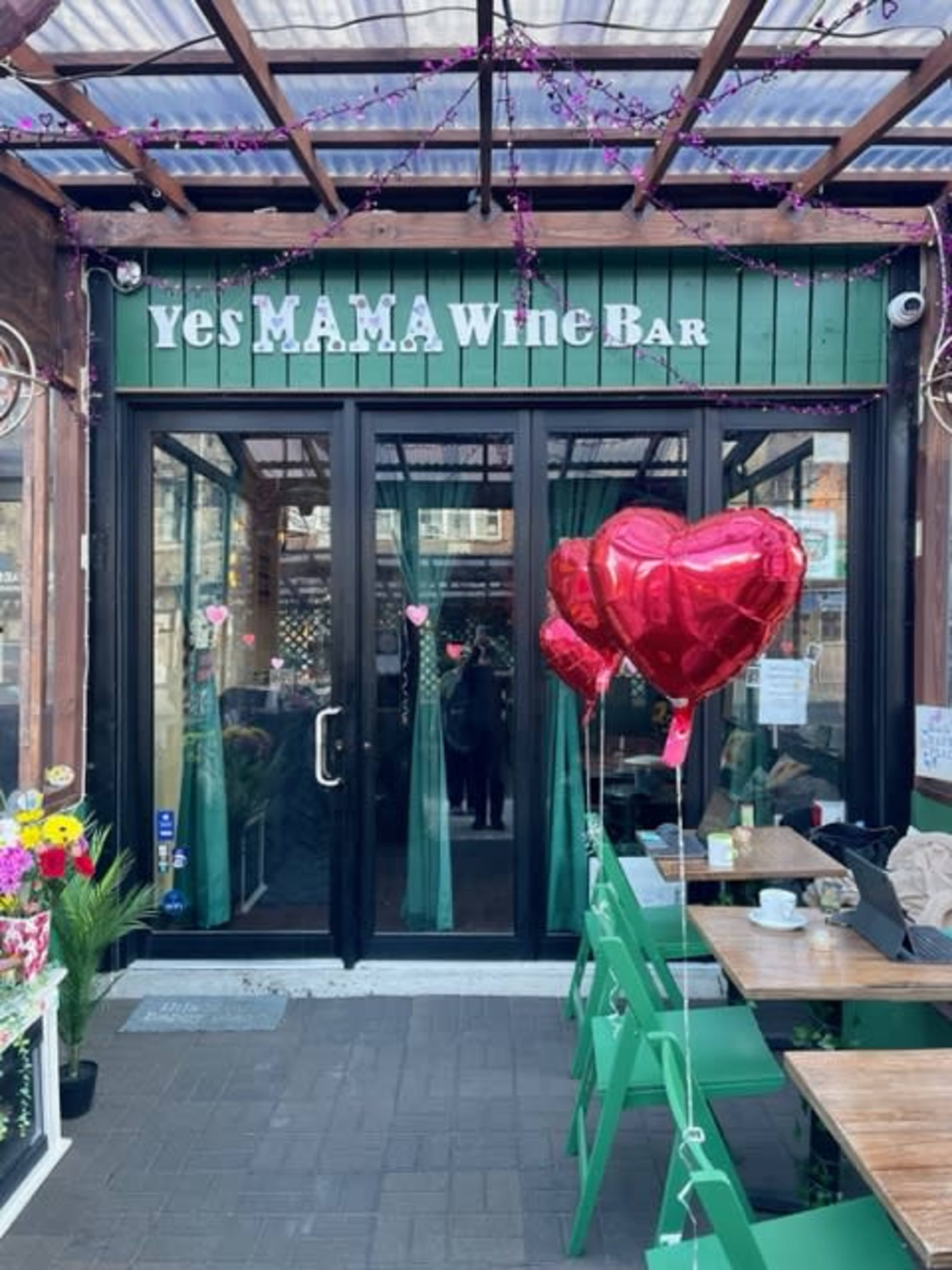 The entrance of "Yes MAMA Wine Bar," featuring large red heart-shaped balloons and a wooden exterior with green accents.