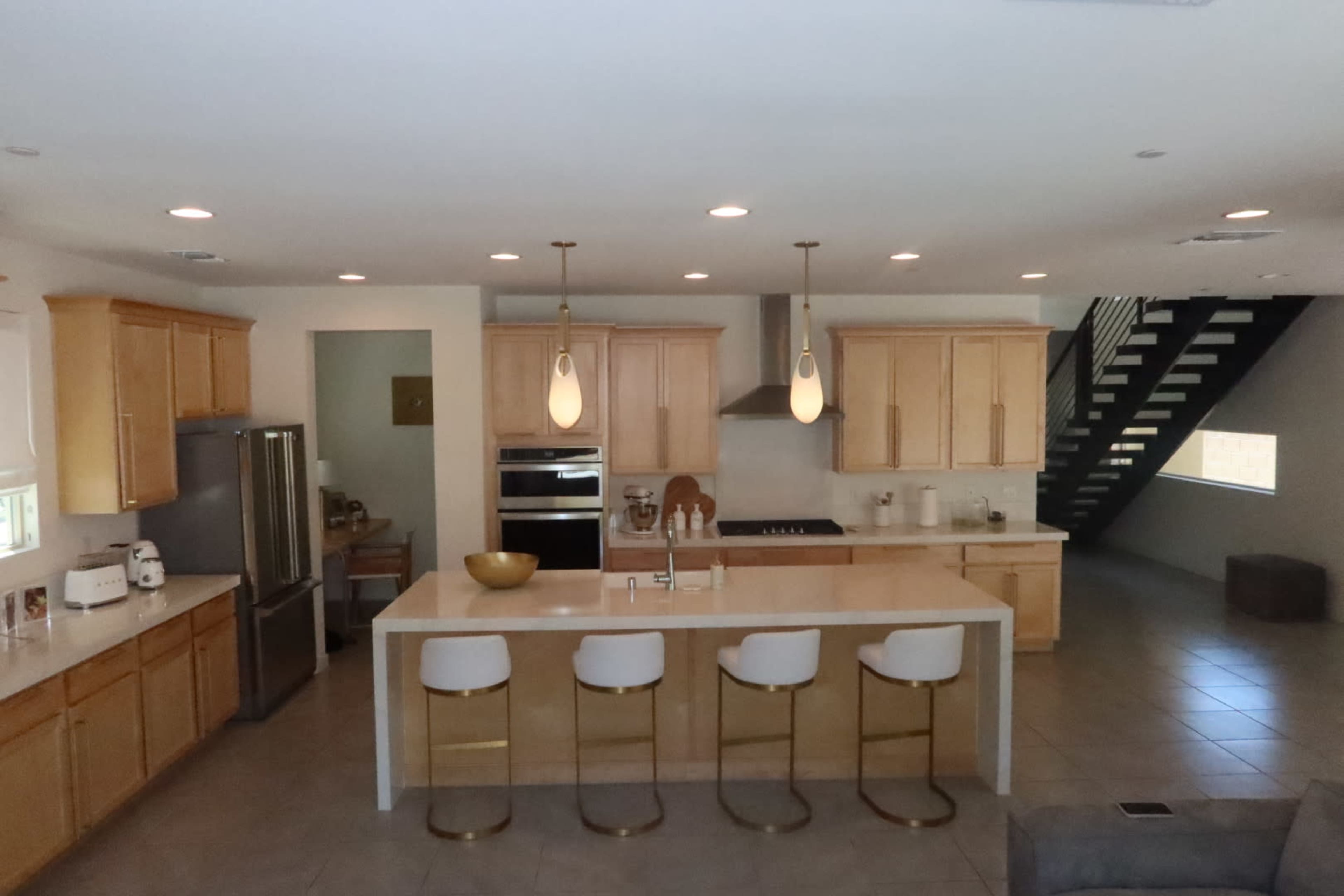 The image shows a modern kitchen with light wood cabinetry, a large island with four white bar stools, and stainless steel appliances.
