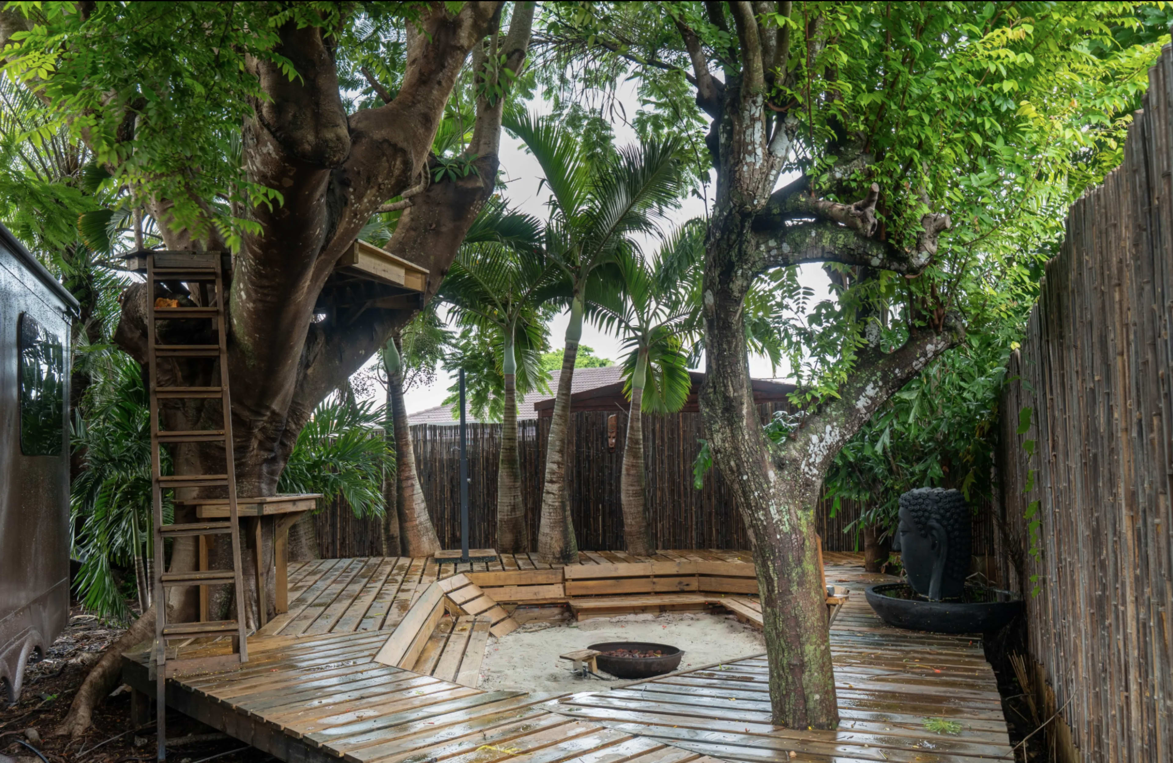 The image shows a wooden deck encircled by trees and lush foliage, with a fire pit in the center and an elevated platform nearby.