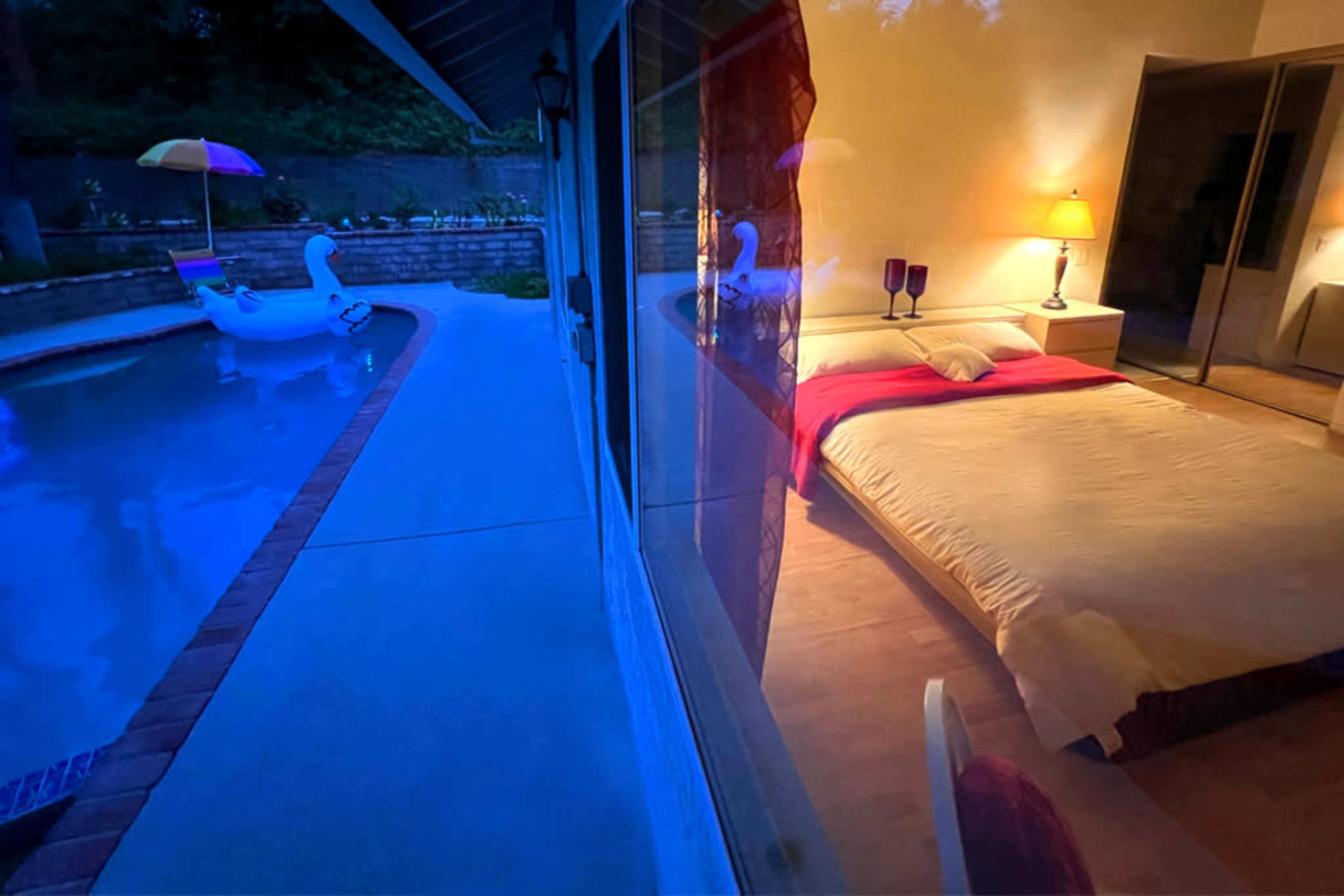 A bedroom with a bed and lamp is reflected in a glass window, overlooking a pool with a swan float and colorful umbrella.