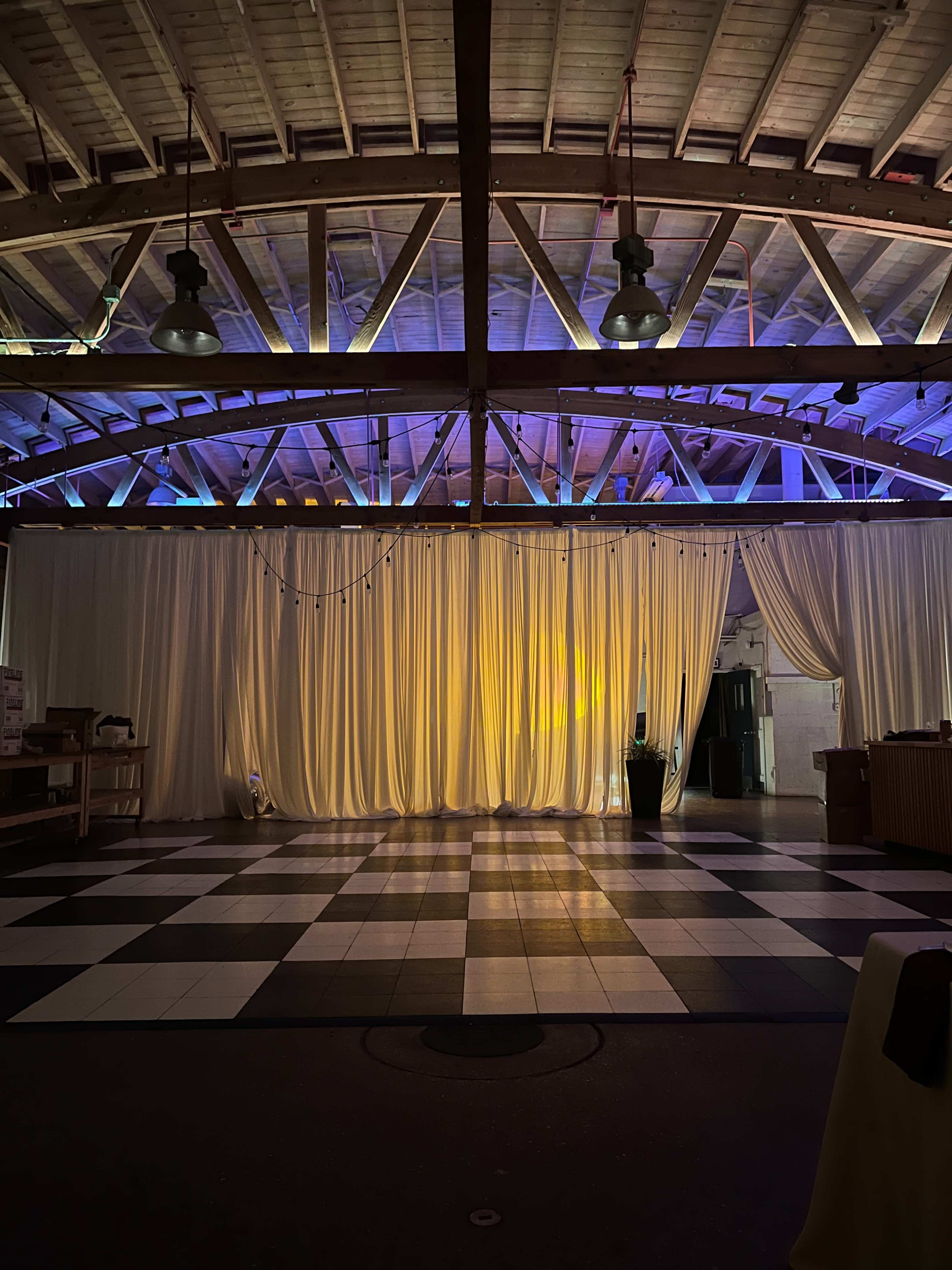 The image shows an event space with a checkered floor, dim lighting, and draped curtains, set under a wooden ceiling with exposed beams.