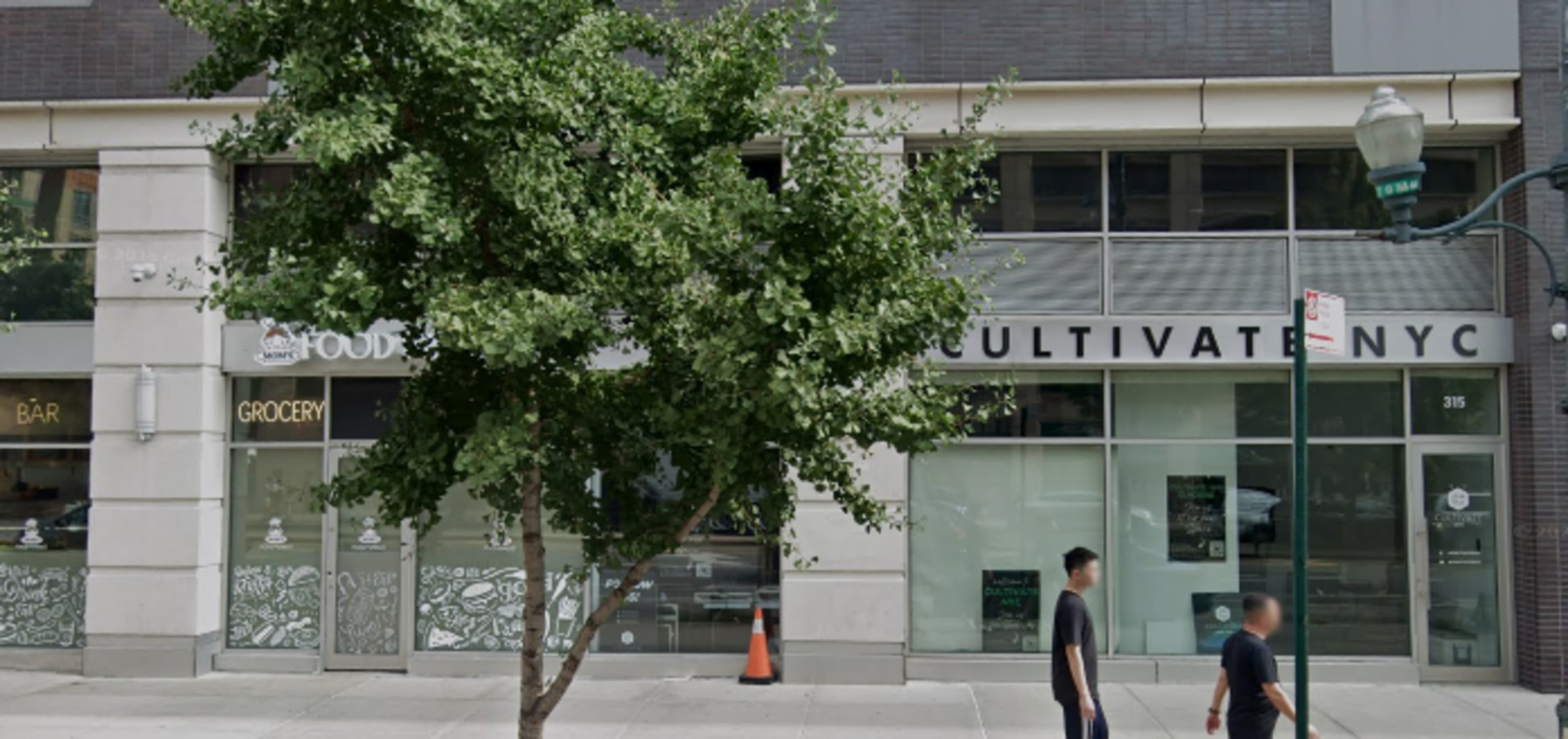 The image shows a commercial building facade in an urban setting with two storefronts, labeled "FOOD" and "CULTIVATE NYC," and a tree in front.