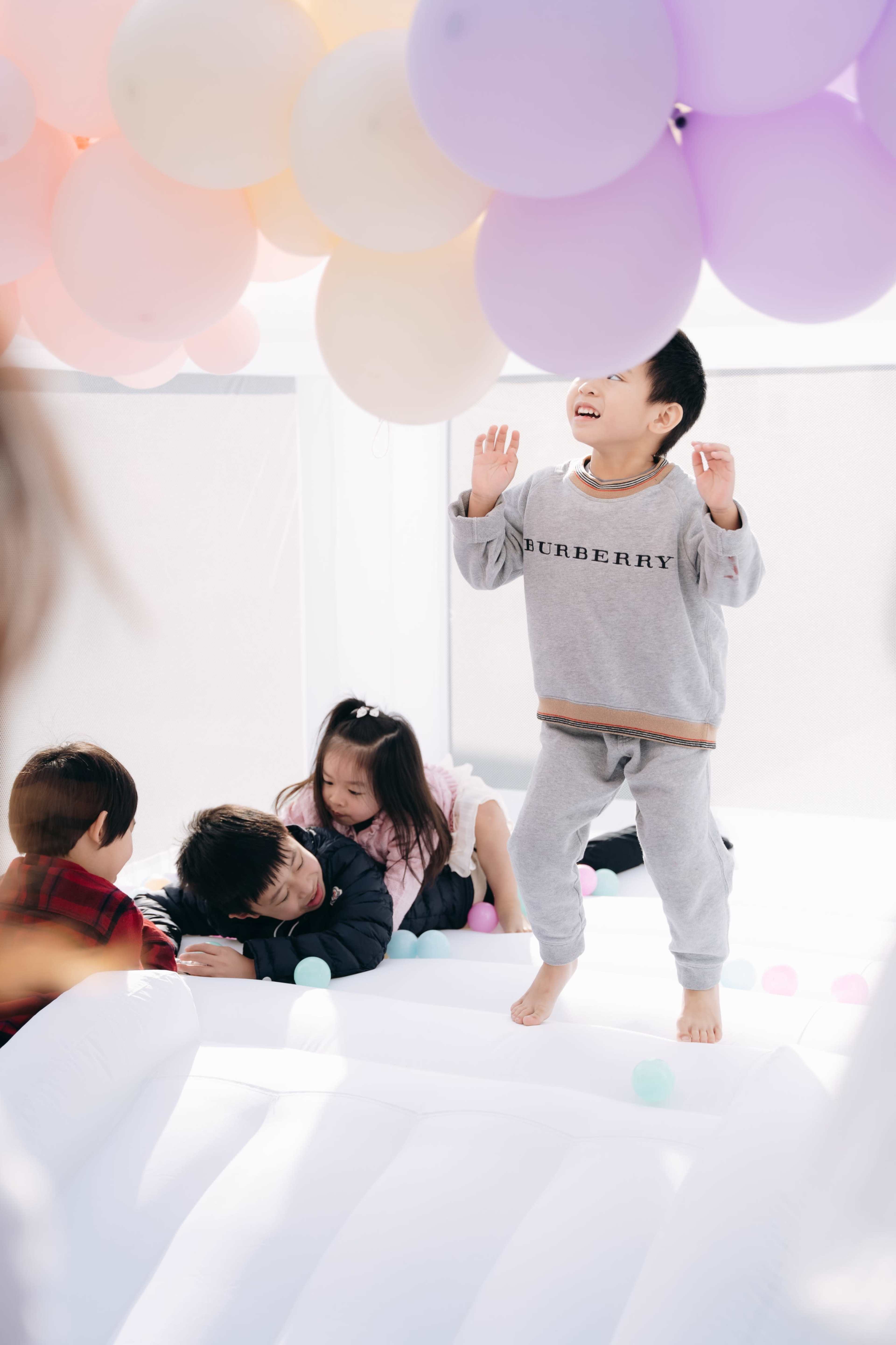 A group of children play and jump on a bouncy surface adorned with colorful balloons overhead.