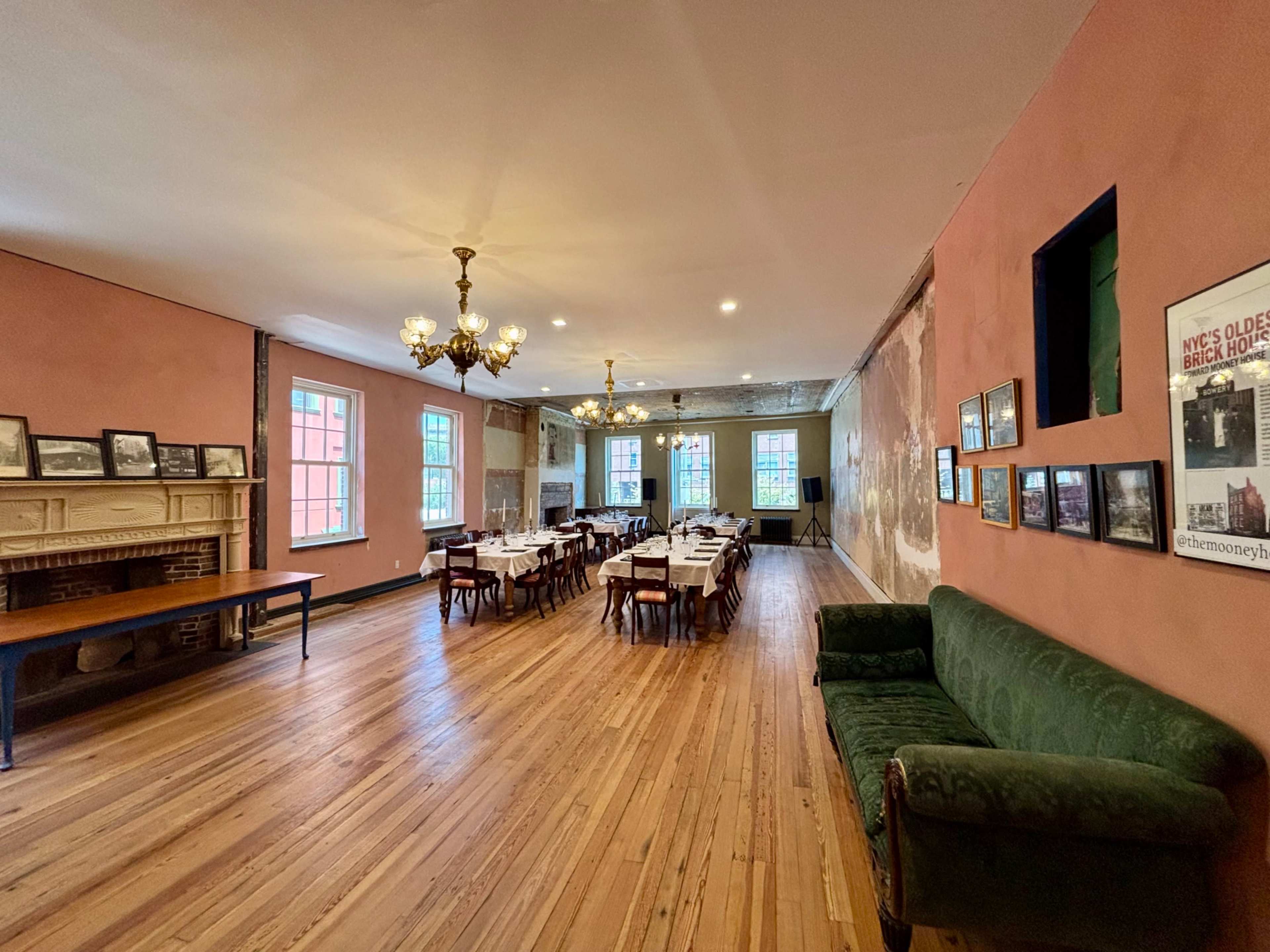 The image shows a spacious dining room with wooden floors, several tables set for guests, a green sofa along one wall, and framed photographs hanging on another wall.