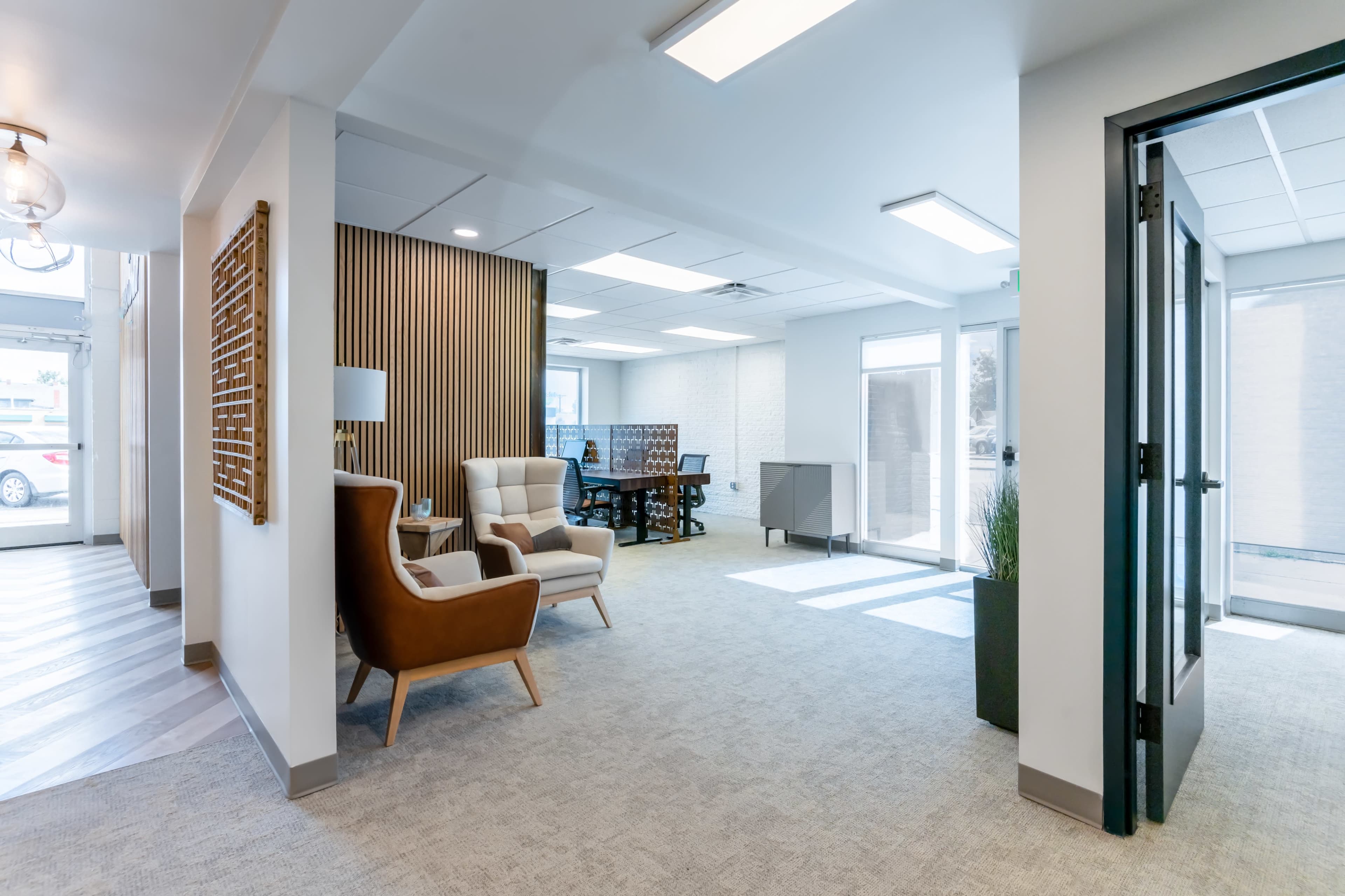A modern office space featuring a seating area with two chairs, a wooden accent wall, and a workspace with a desk in the background.
