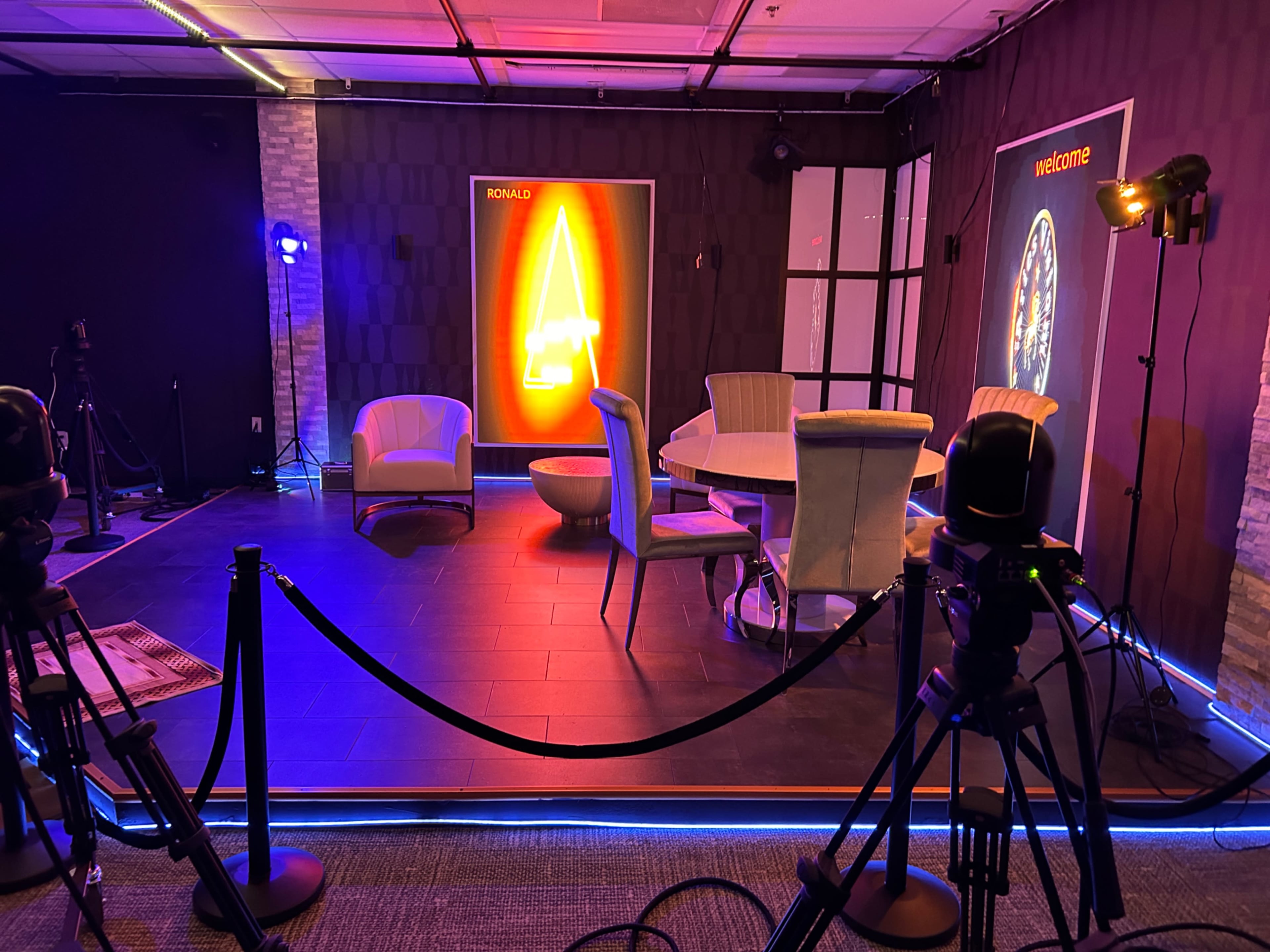 The image shows a dimly lit room set up for a presentation, featuring a round table surrounded by chairs, with colorful lighting and a backdrop that includes a neon sign.
