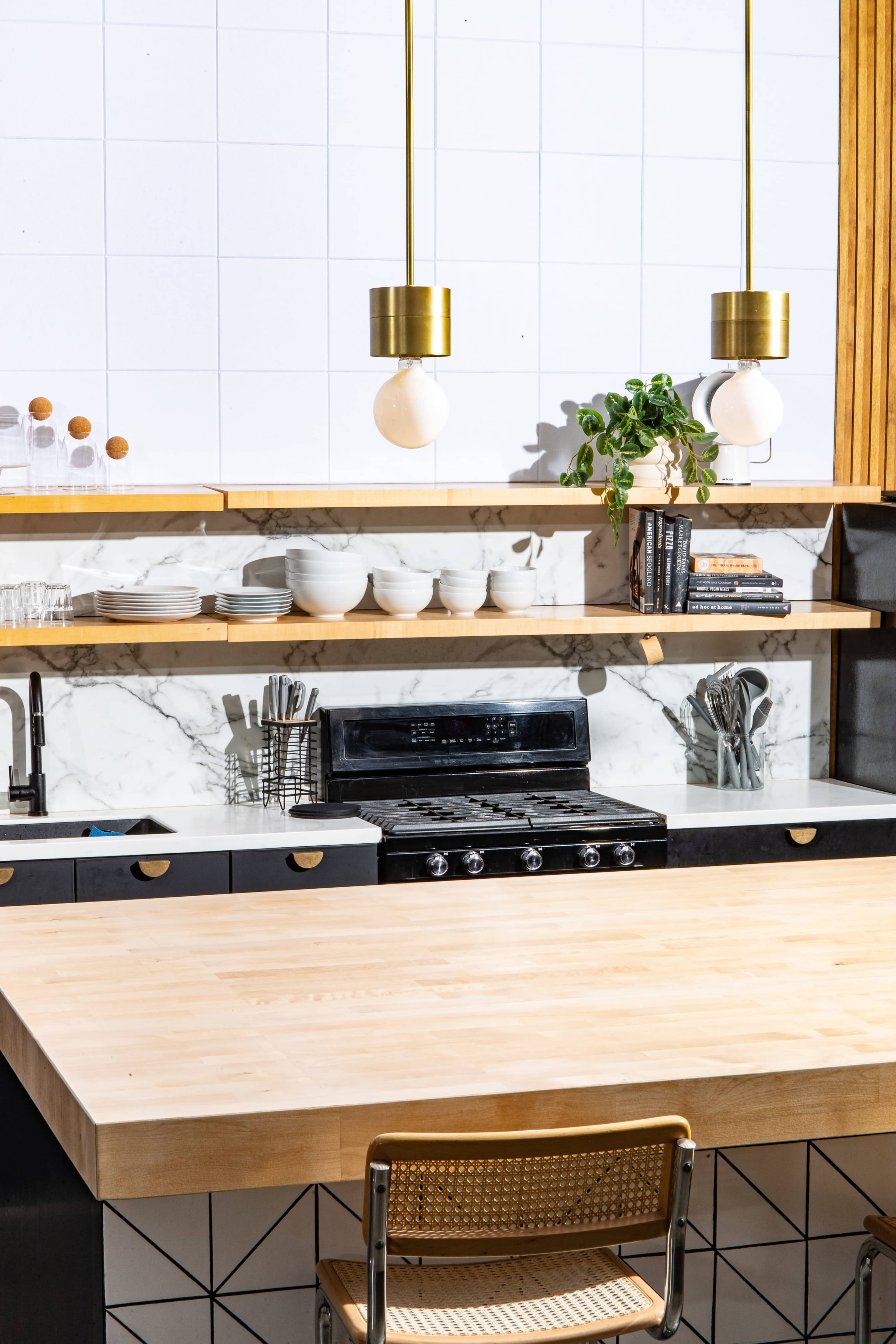 A modern kitchen features a wooden countertop, brass pendant lights, marble backsplashes, and open shelves with dishware and utensils.
