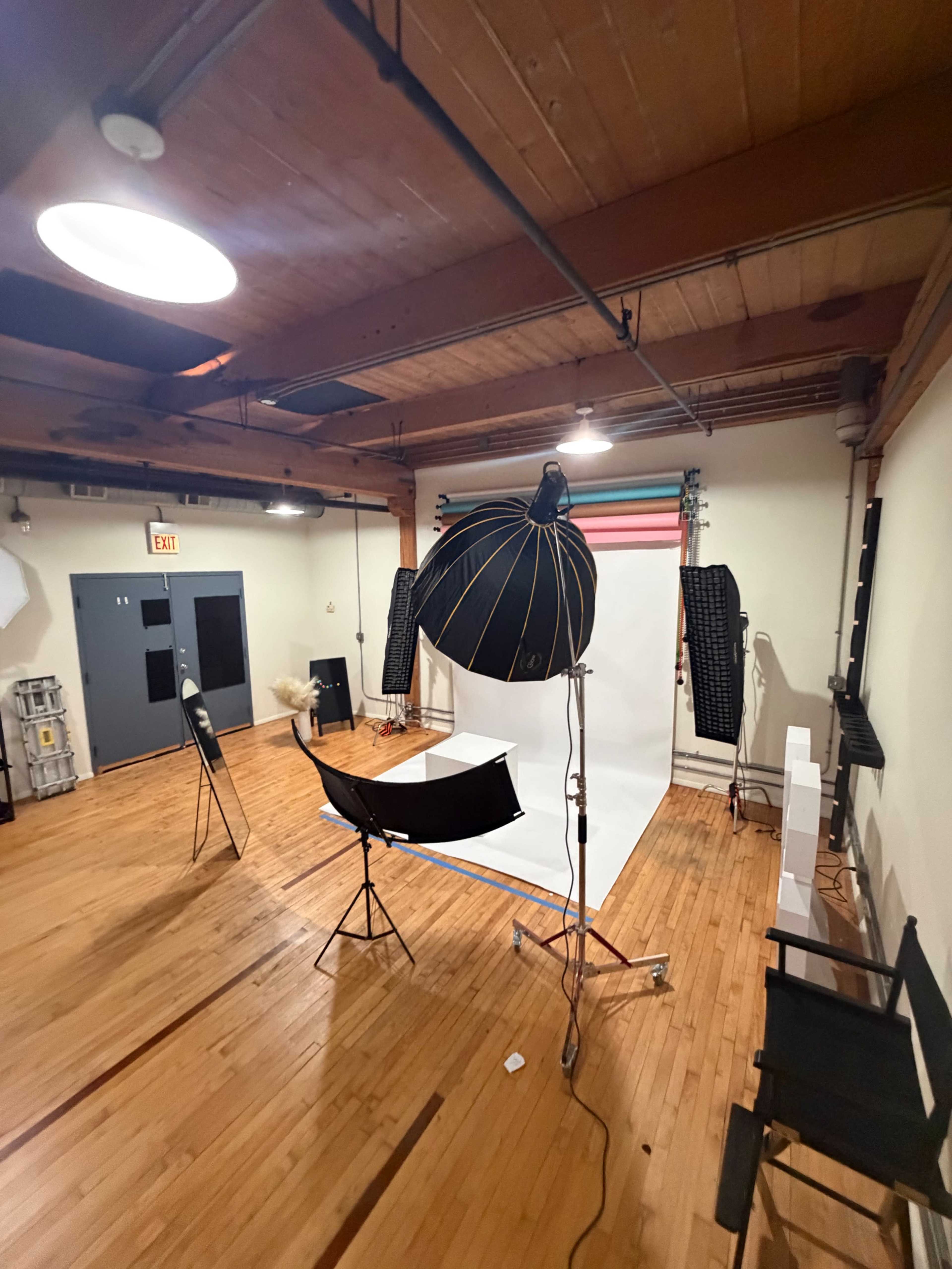 The image shows a photography studio with a large softbox, two light stands, a backdrop of colored stripes, and several chairs arranged in the space.