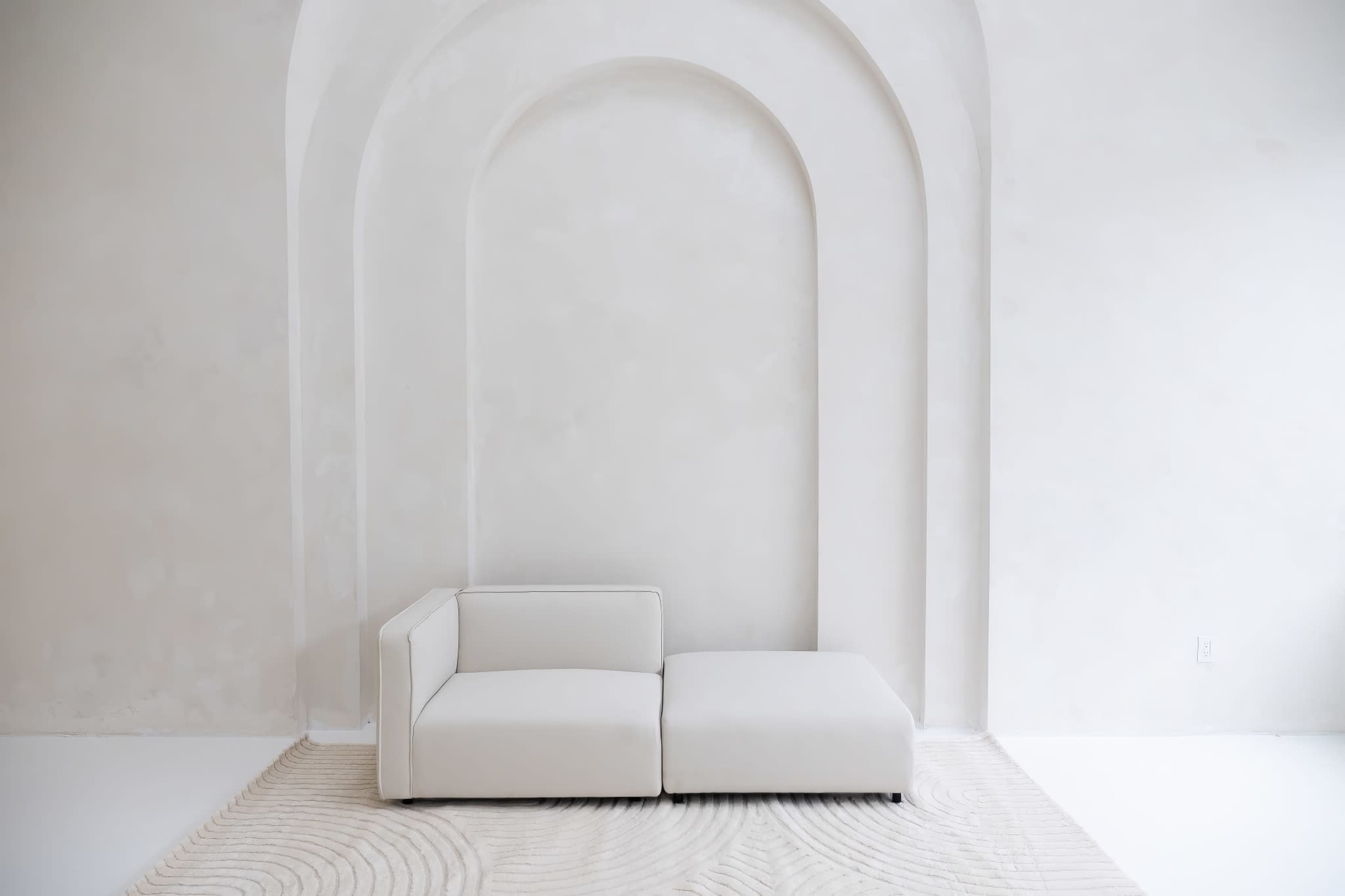 A modern, white sectional sofa is positioned against a light, arch-shaped wall in a minimalist room with a textured rug on the floor.
