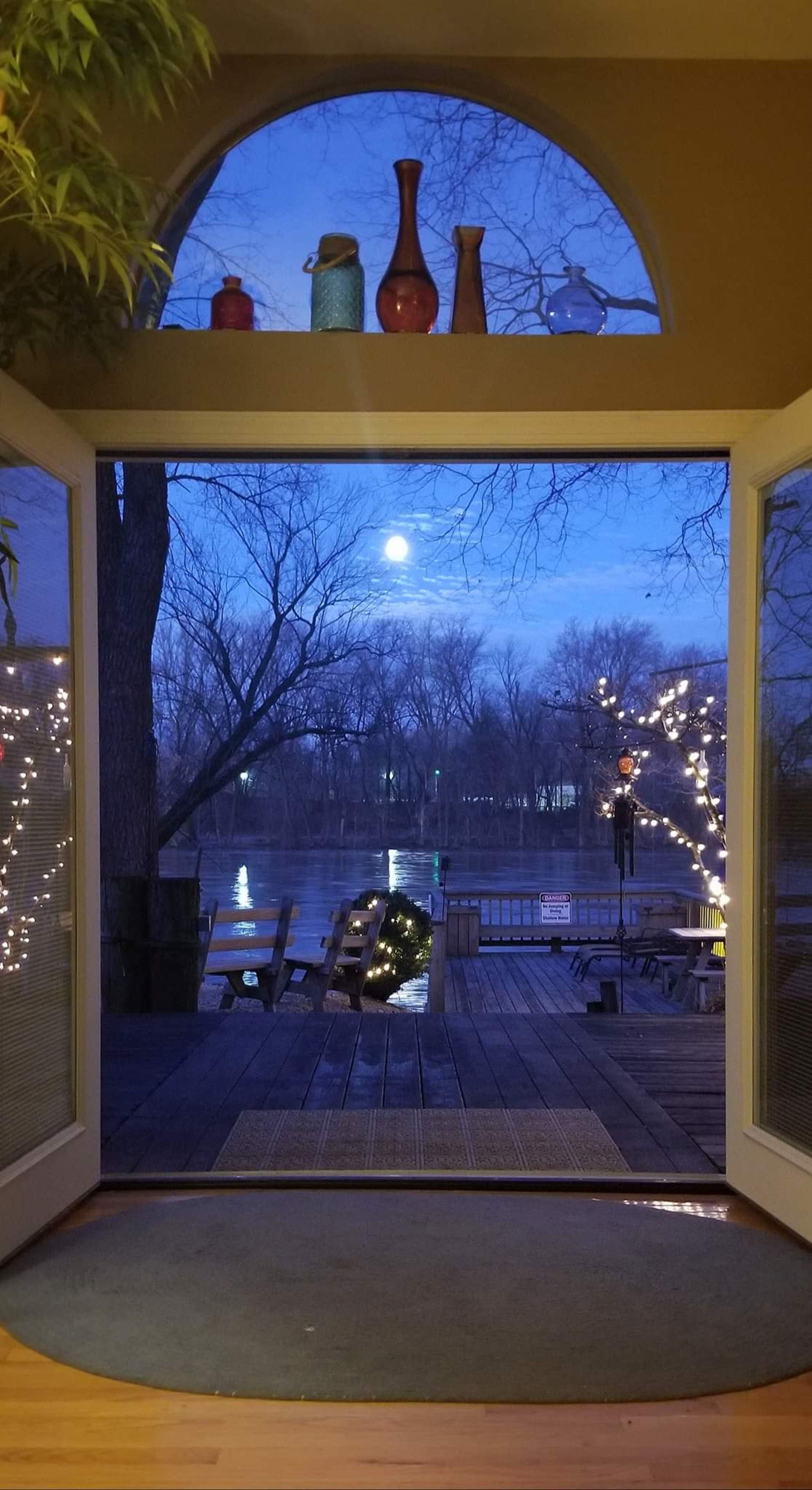 A view of a river with trees and a deck illuminated by lights, framed by open double doors under a moonlit sky.