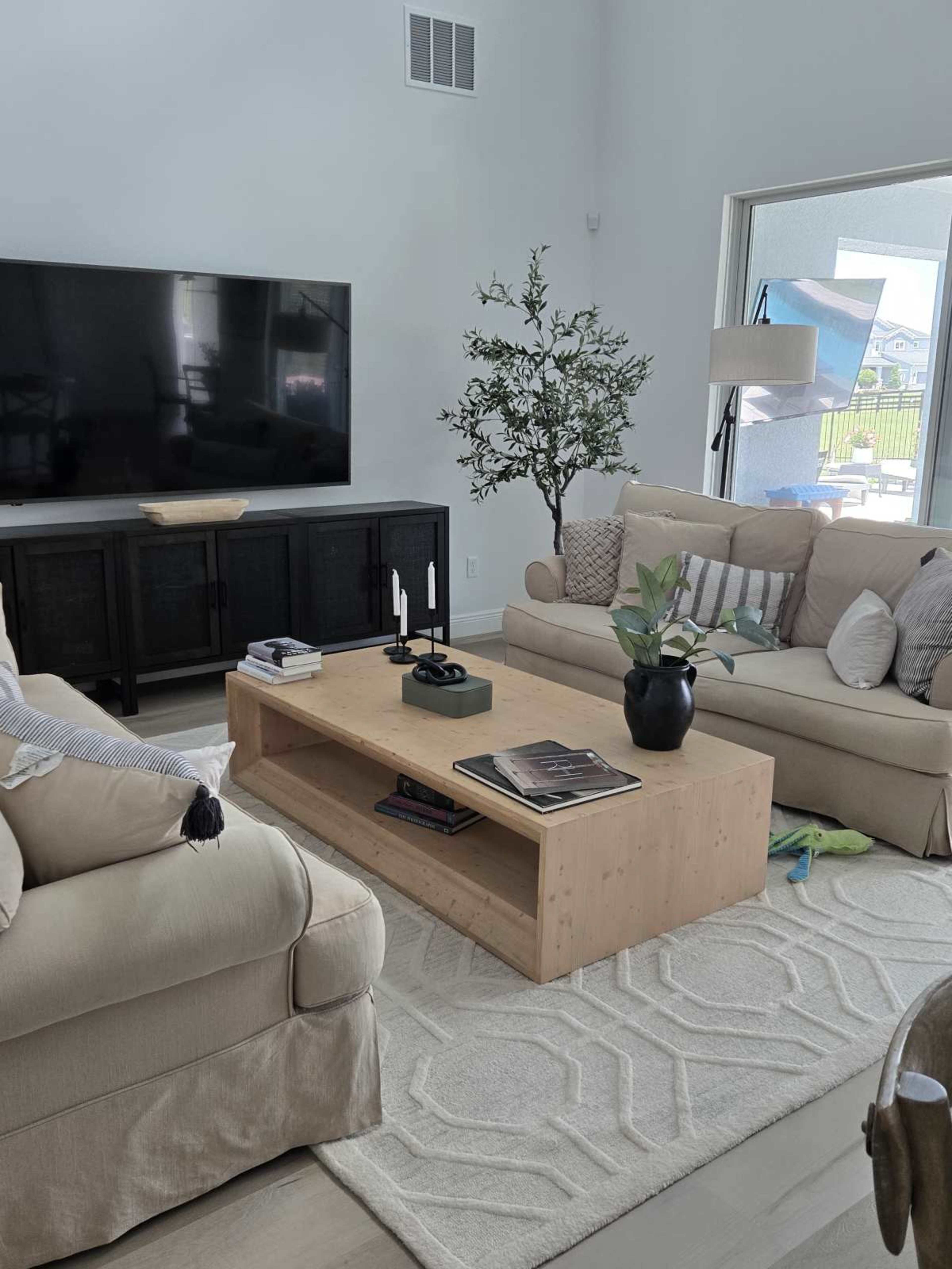 The image shows a modern living room with a large television on the wall, two beige couches, a wooden coffee table, and decorative plants.