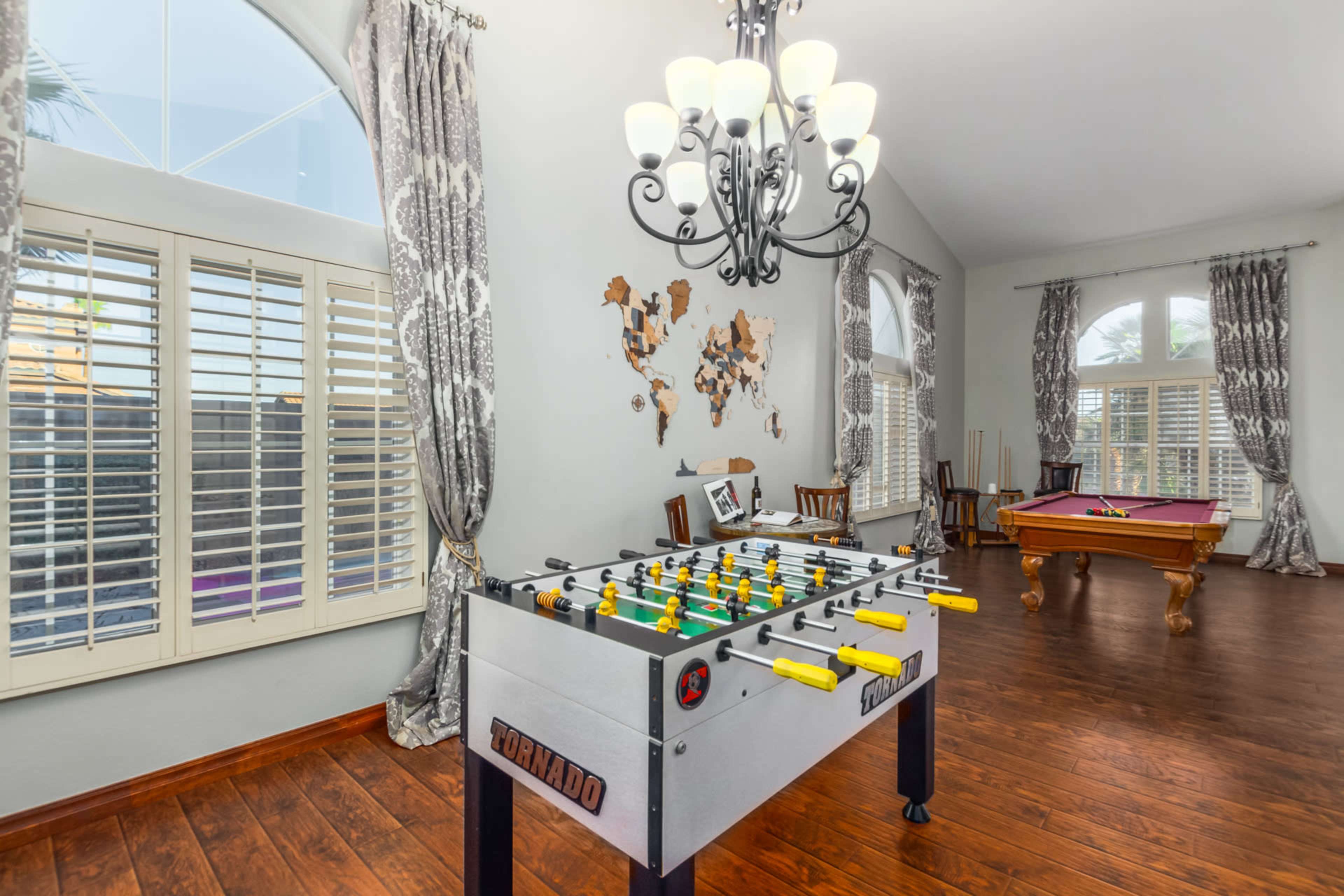 The room features a foosball table in the foreground, with a billiards table and large windows in the background.