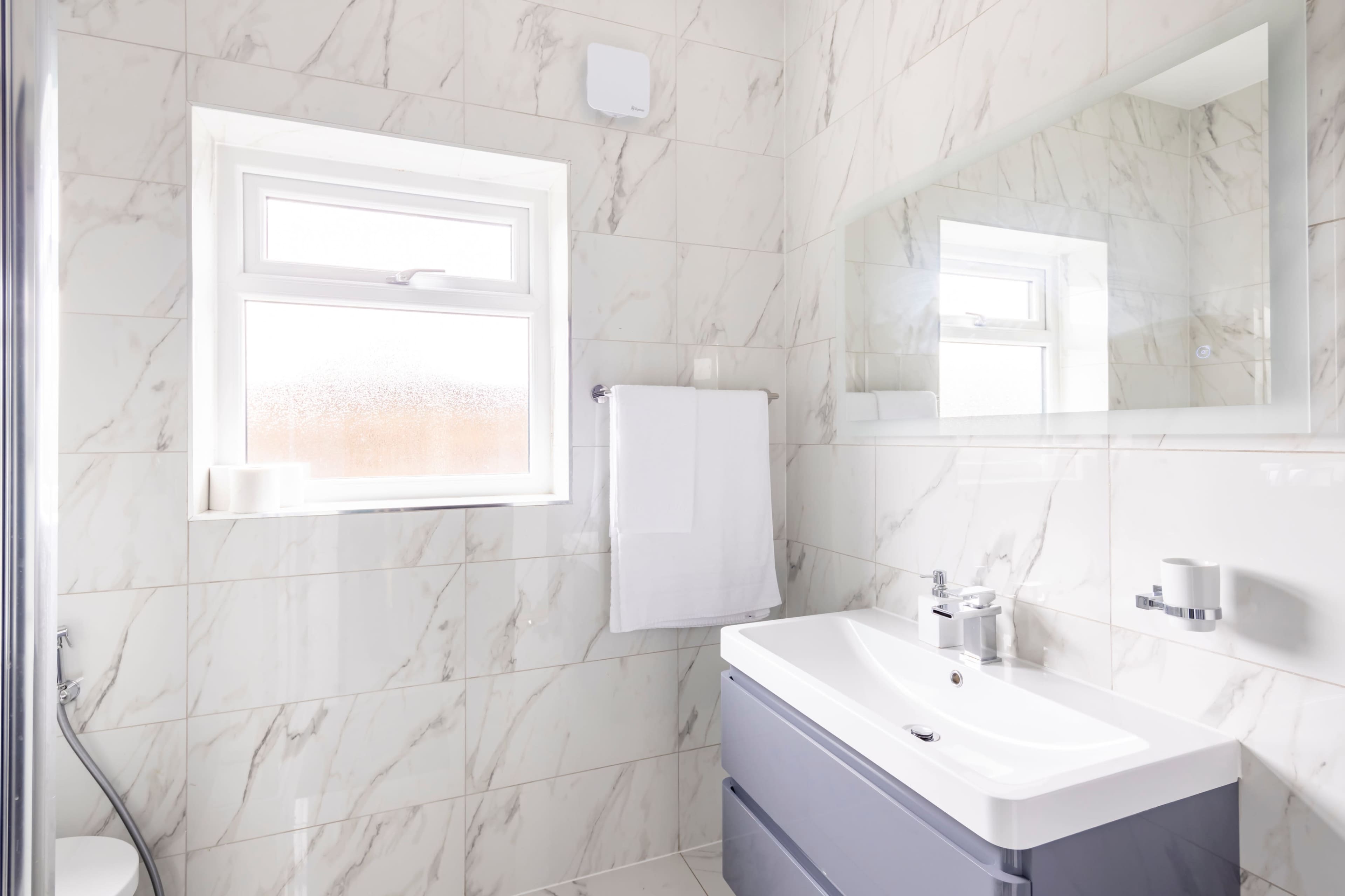 The image shows a modern bathroom featuring a marble-tiled wall, a white sink with a gray cabinet, a mirror, a towel rack, and a frosted window.