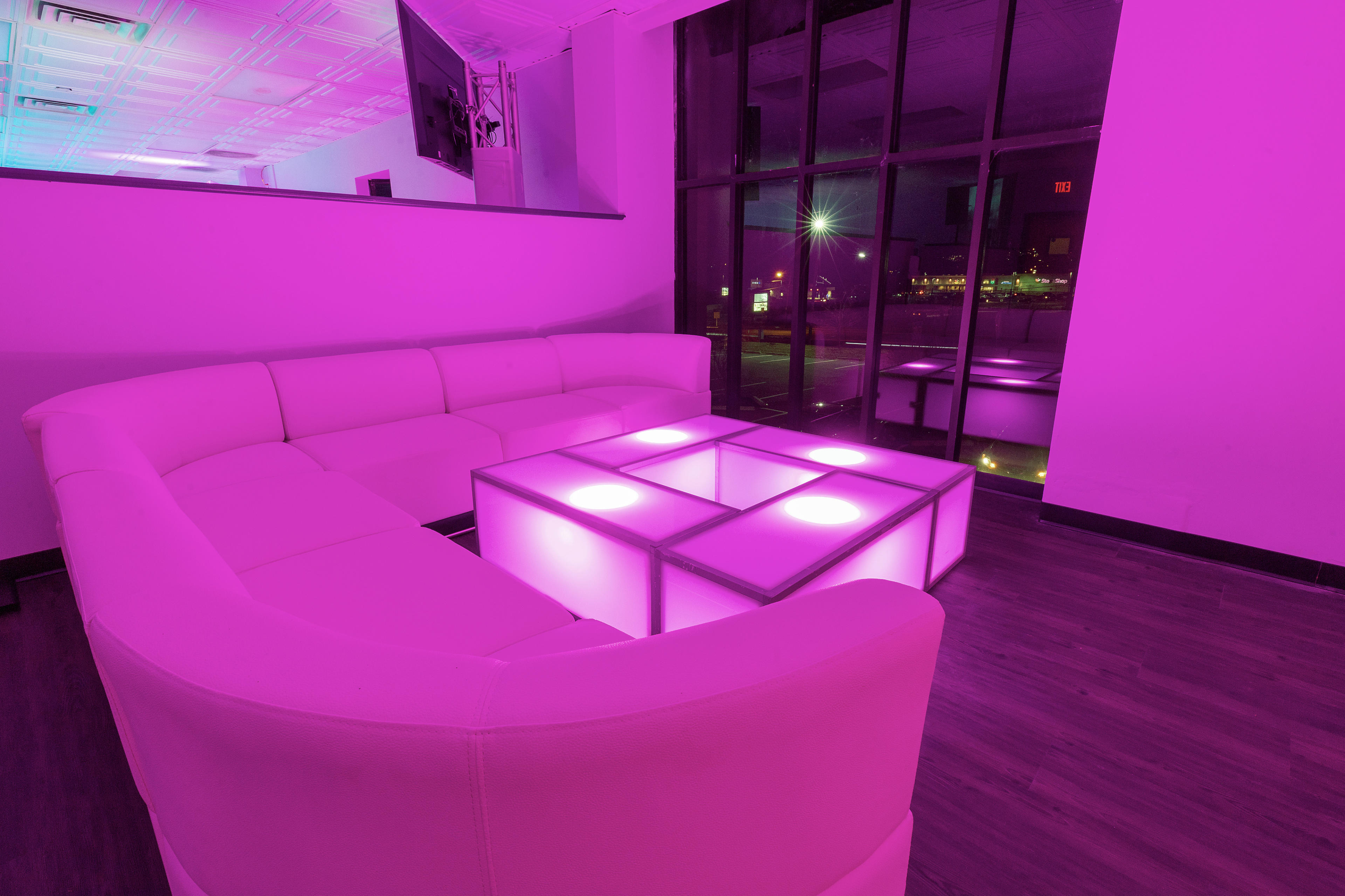 The image shows a modern lounge area featuring a white sectional sofa and a lit square coffee table in a softly illuminated pink space.