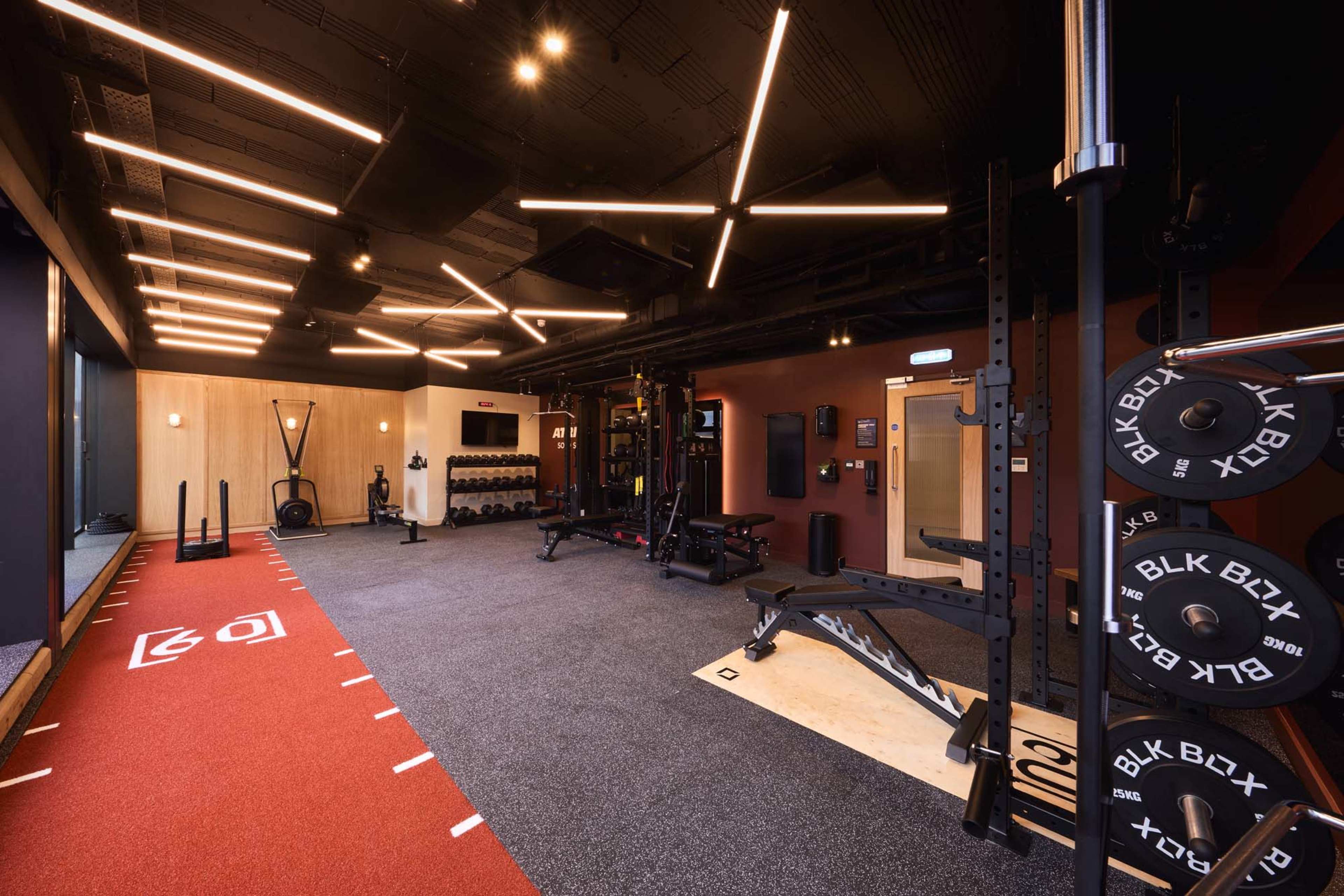 The image shows a modern gym interior featuring various workout equipment, including weights, benches, and training stations, with overhead lighting and a designated exercise area.