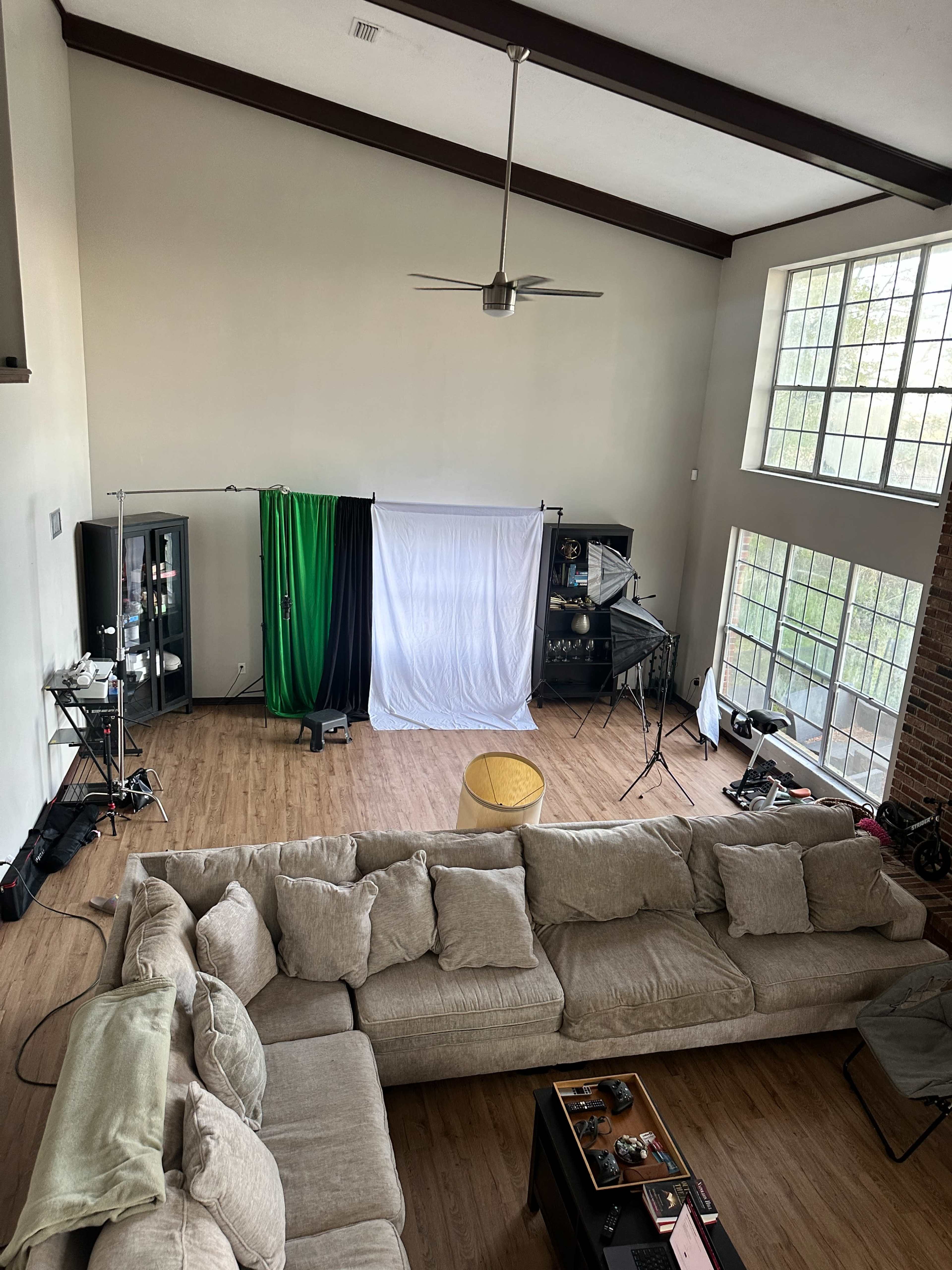 A spacious living room features a large sectional sofa, a coffee table with items on it, and a backdrop setup for photography in the corner, illuminated by natural light from large windows.