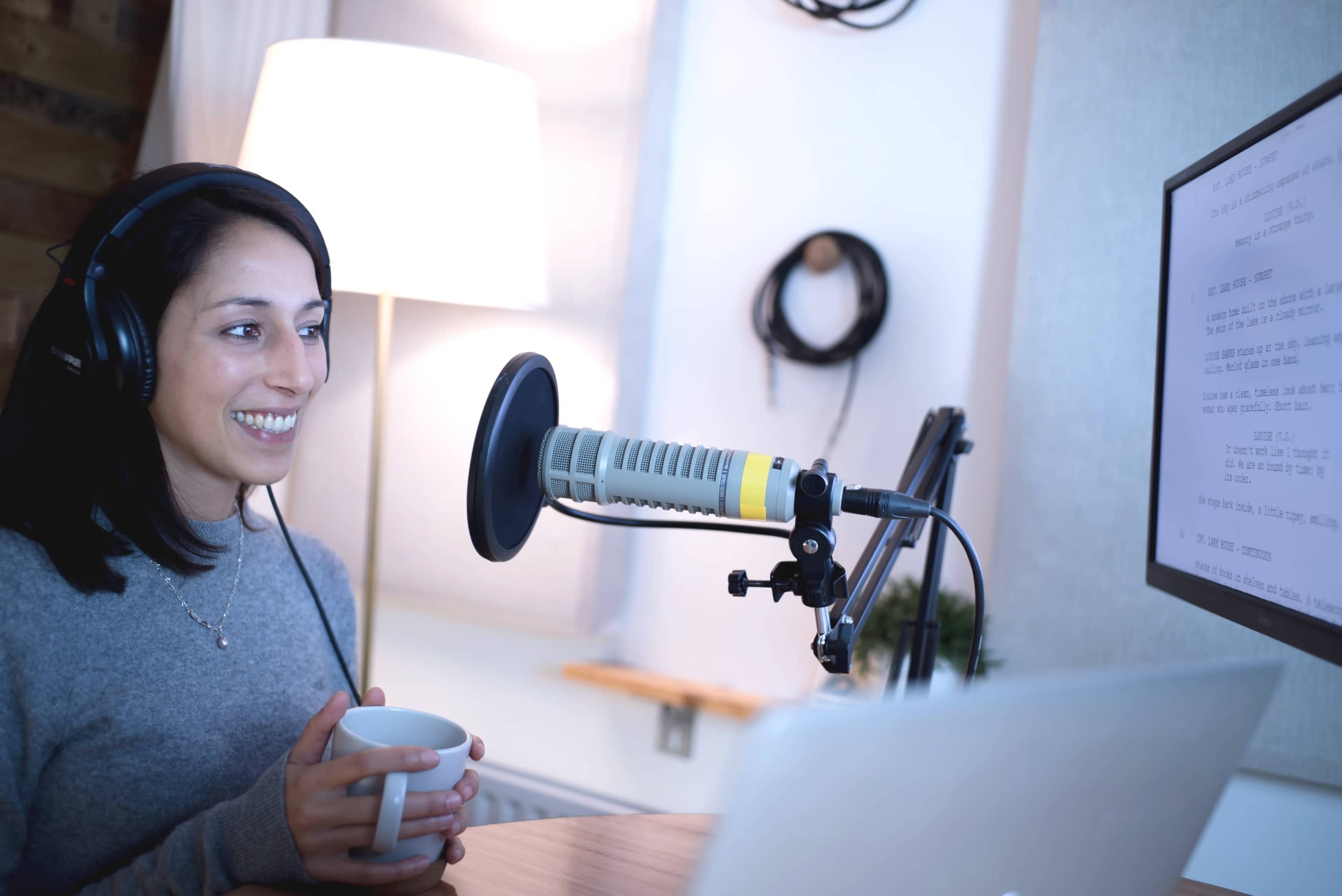 A woman wearing headphones sits at a desk with a microphone and a laptop, holding a cup, in a well-lit recording studio.