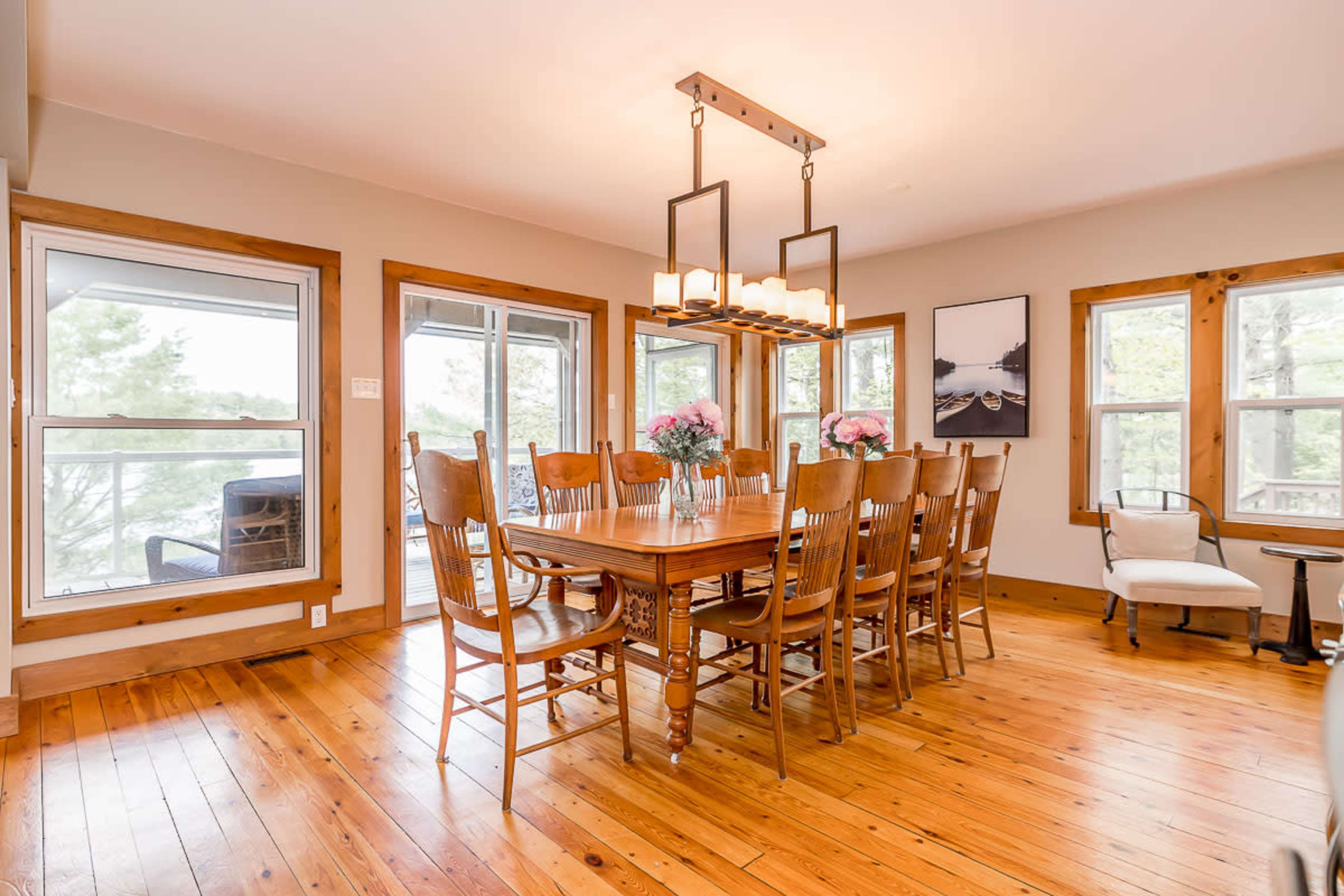 A spacious dining area features a large wooden table surrounded by ten chairs, with large windows allowing natural light and a view of the outdoors.
