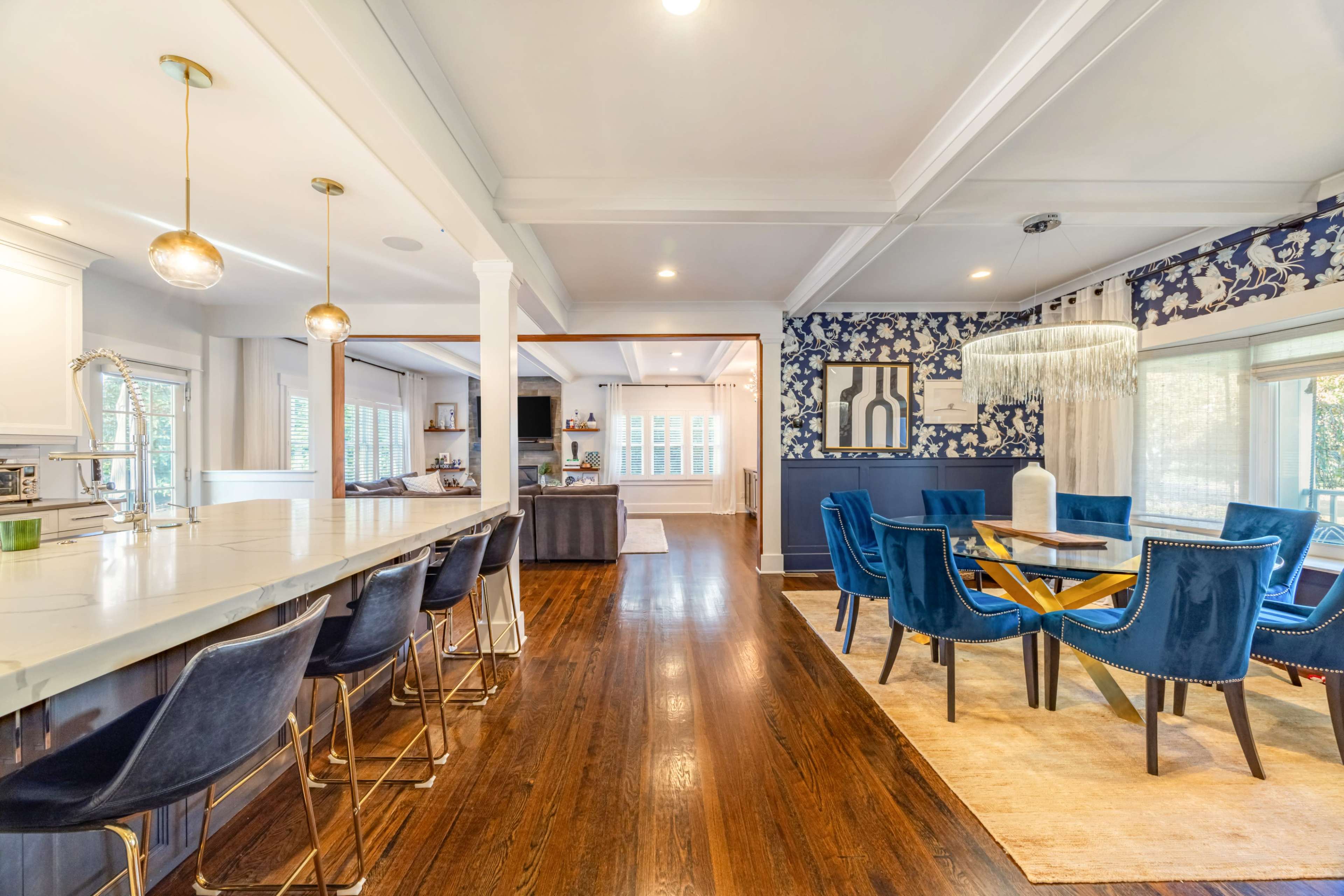 The image shows a modern open-concept kitchen and dining area with a bar featuring high stools, a dining table surrounded by blue chairs, and decorative wallpaper.