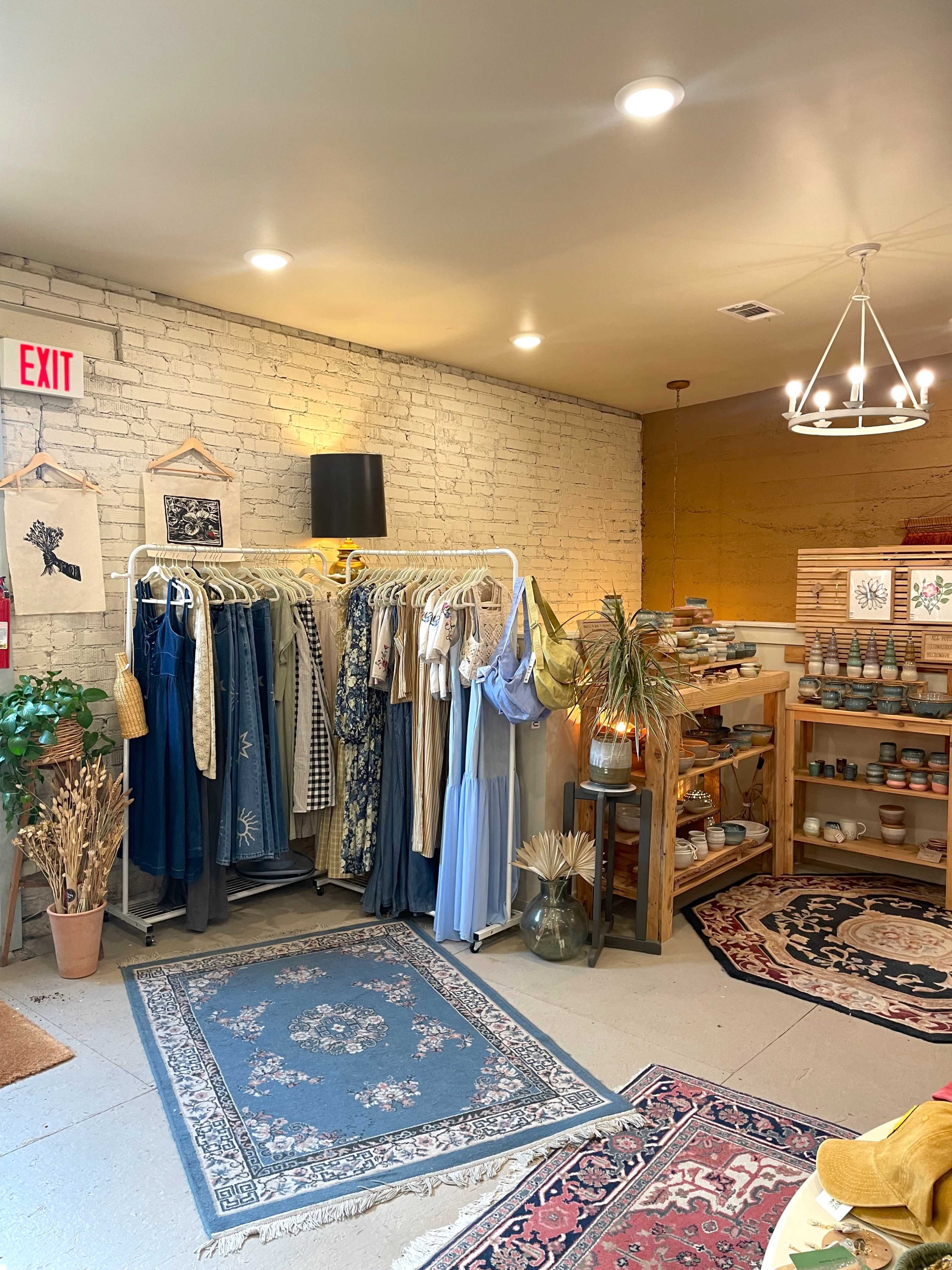 The image shows a boutique interior with a clothing rack displaying various garments, a wooden shelf filled with products, and decorative rugs on the floor.