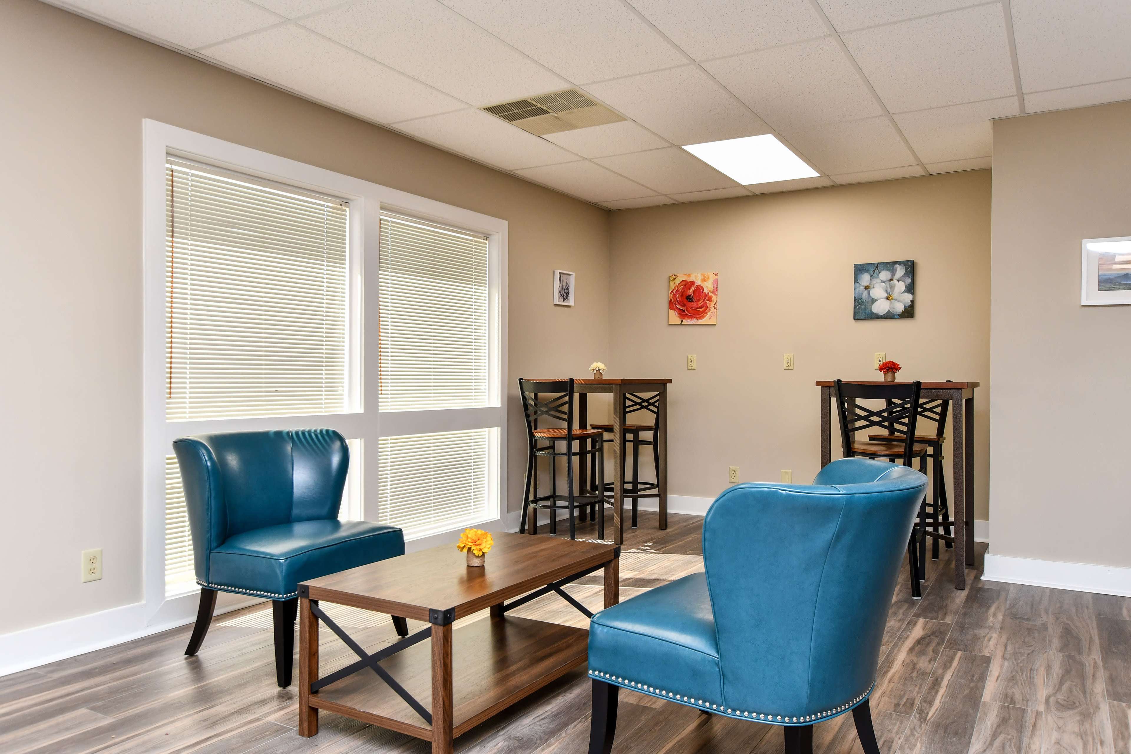 A small lounge area featuring two blue chairs, a wooden coffee table, and high tables with chairs against the walls.